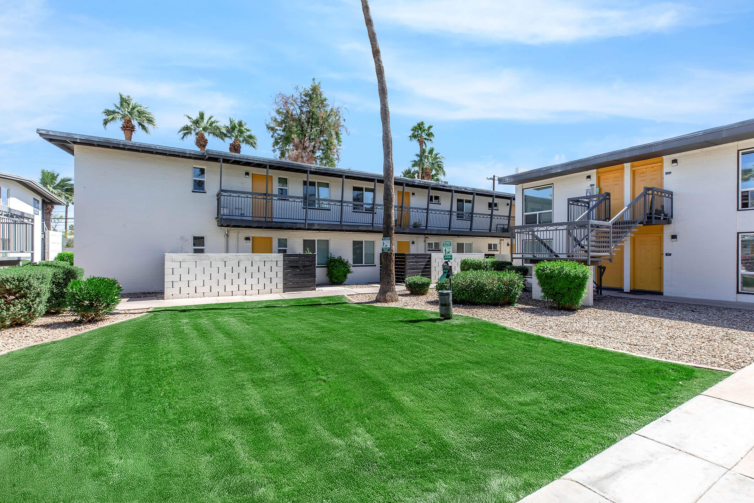 A well-maintained apartment complex with two buildings and a landscaped green lawn. Palm trees provide shade in the background, and the area is paved with concrete. The buildings feature balconies and a modern design, creating a welcoming atmosphere in a sunny environment.