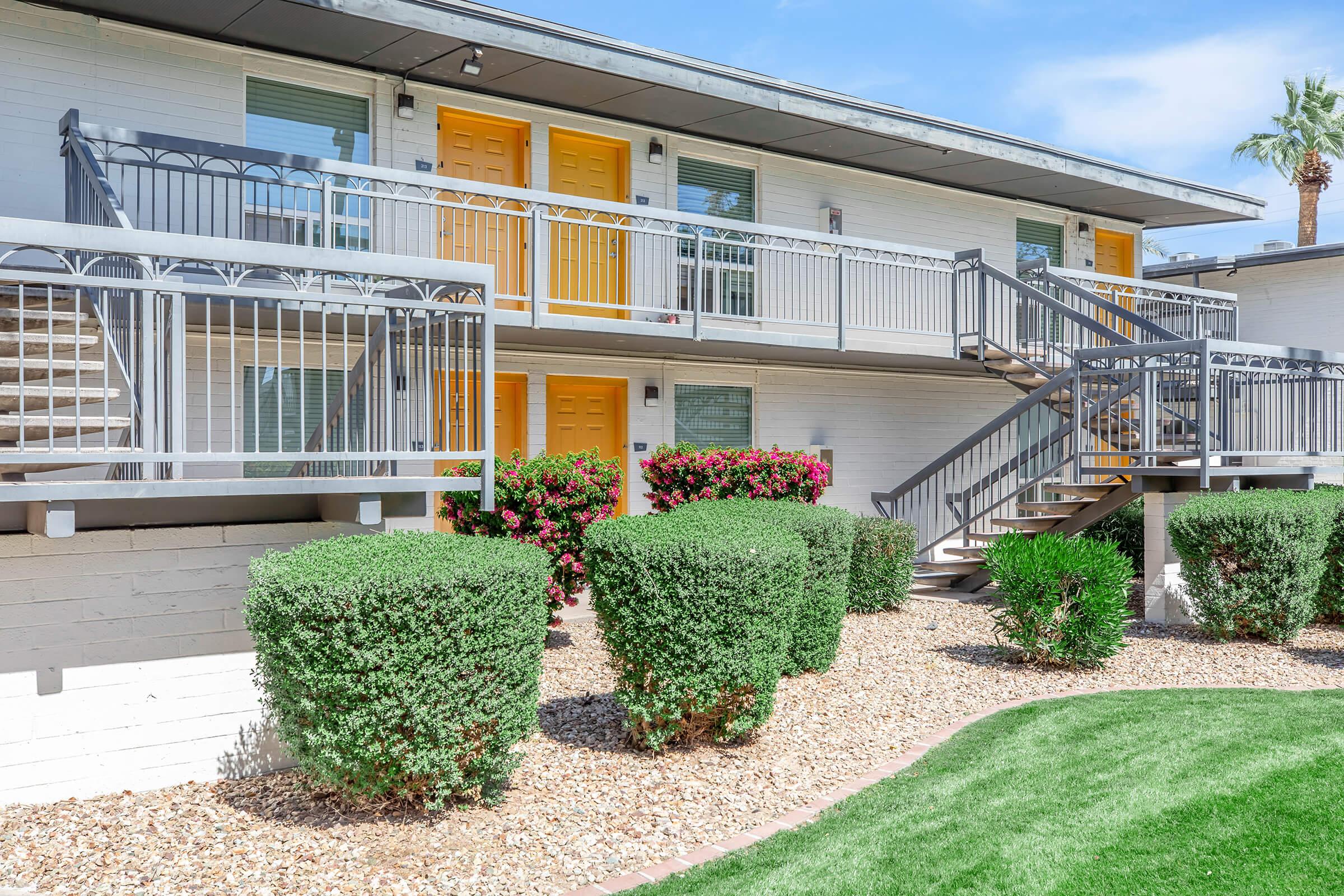 Two-story apartment building with bright yellow doors and white walls. The building features a balcony with a railing, surrounded by neatly trimmed shrubs and a patch of green grass. There are palm trees in the background and a clear blue sky above.