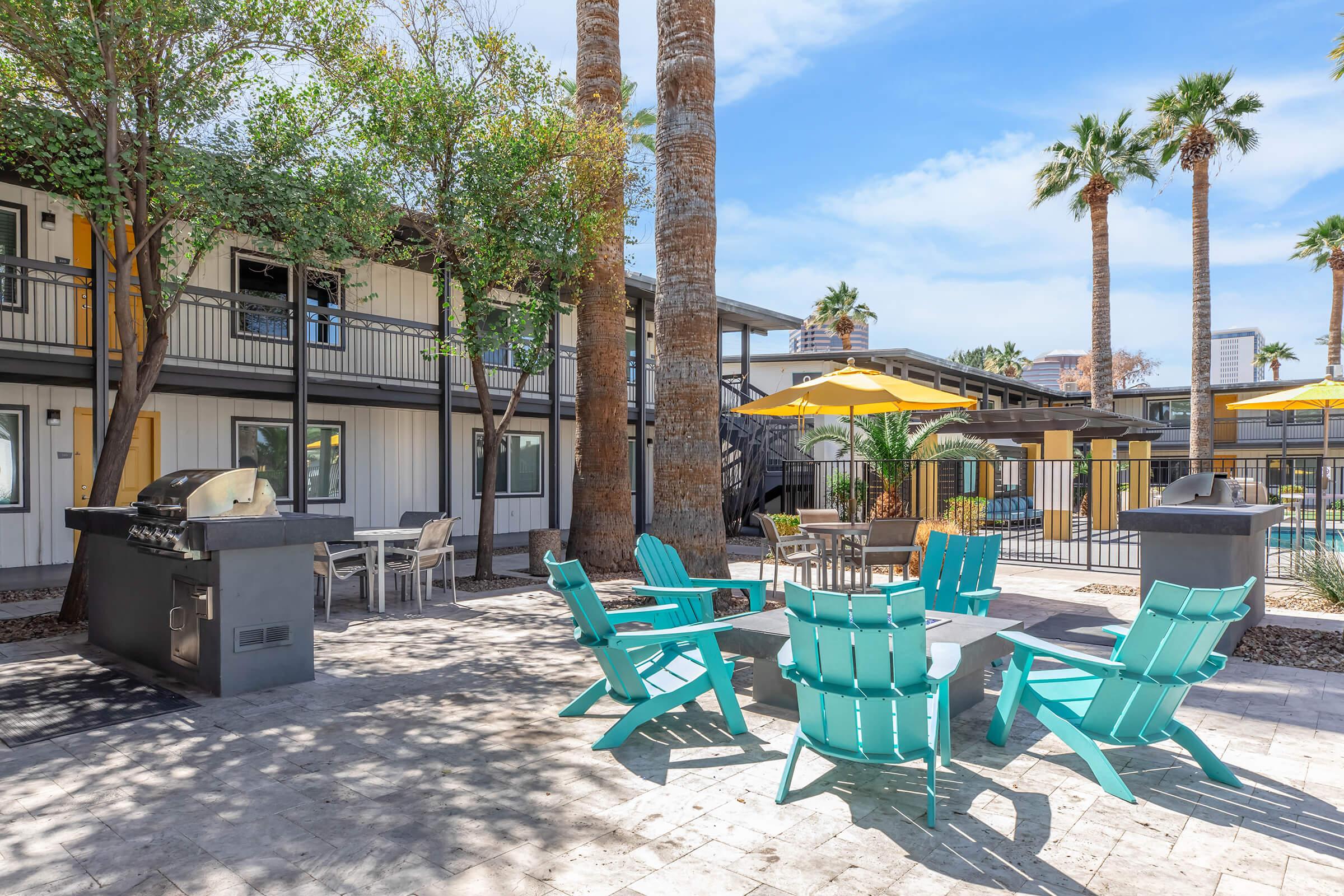 Outdoor patio area featuring turquoise adirondack chairs, a barbecue grill, and a dining table under a yellow umbrella. Surrounded by palm trees and a swimming pool, the space is designed for relaxation and social gatherings. Modern buildings are visible in the background, adding to the inviting atmosphere.