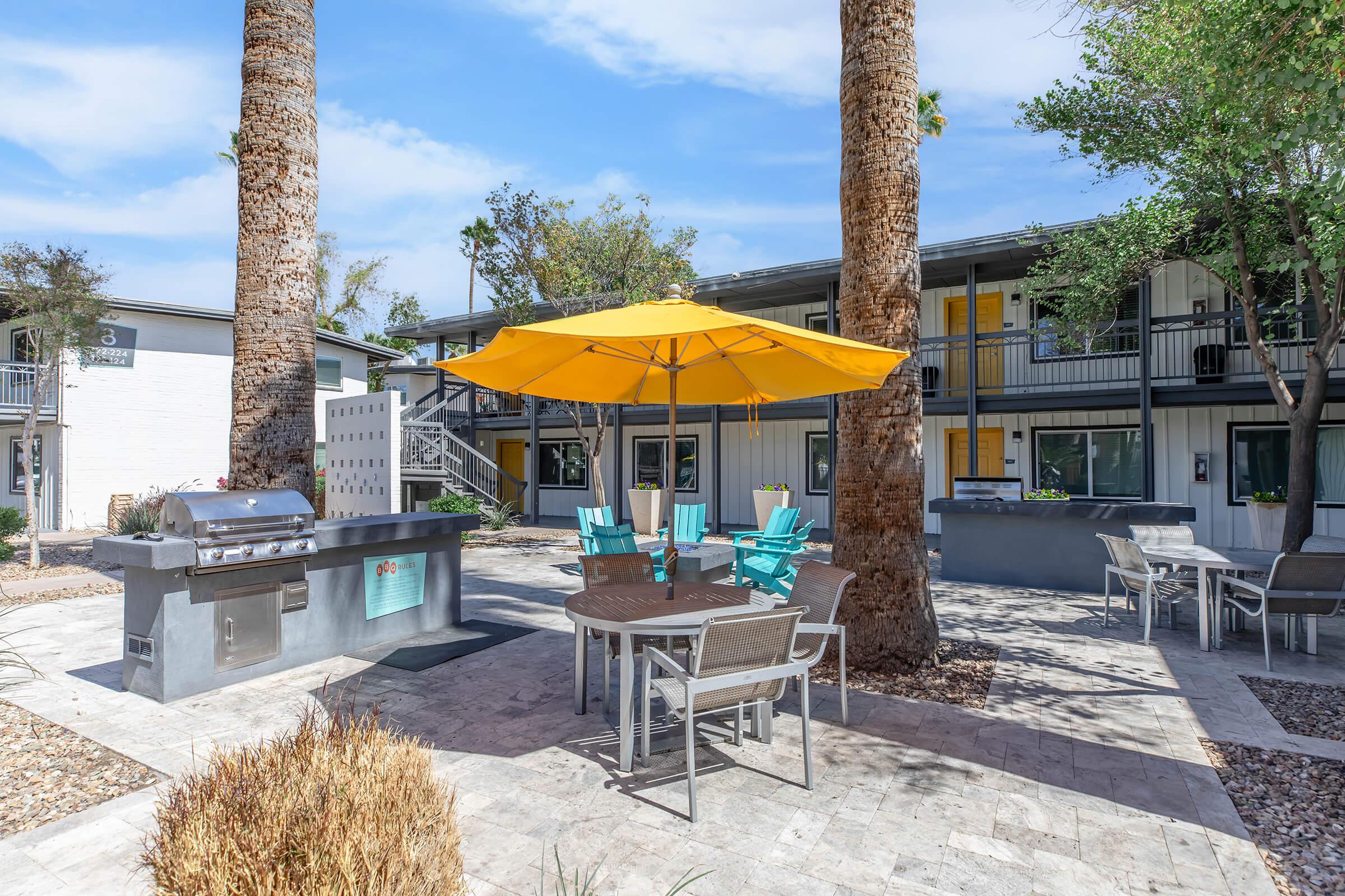 A courtyard at an apartment complex featuring a barbecue grill, a dining table with chairs, and a large yellow umbrella for shade. Surrounding the area are palm trees and modern buildings with balconies, under a clear blue sky.