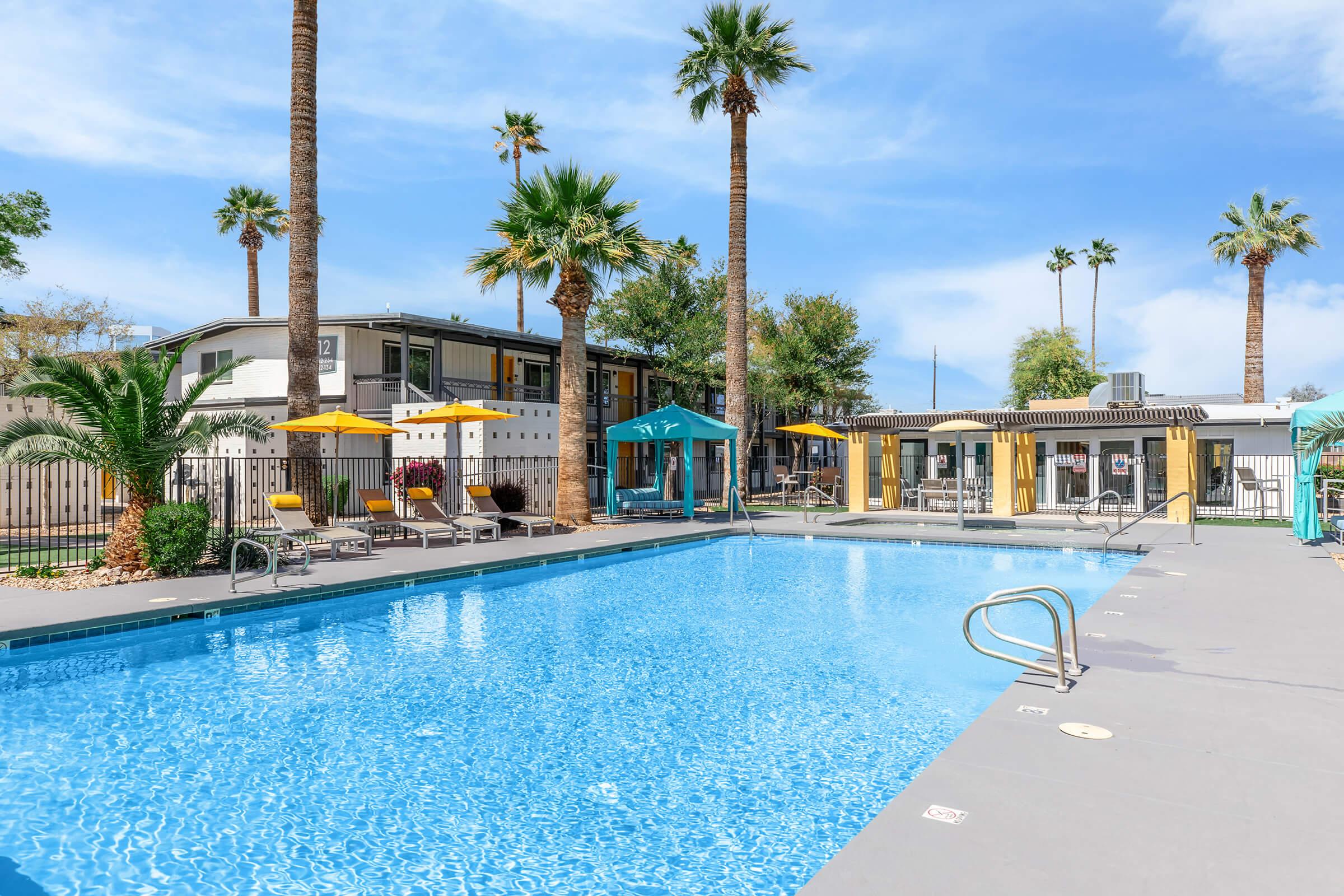A bright and inviting outdoor swimming pool surrounded by palm trees and lounge chairs. Colorful umbrellas provide shade, and a nearby building is visible in the background, creating a relaxing atmosphere. The scene is set under a clear blue sky.