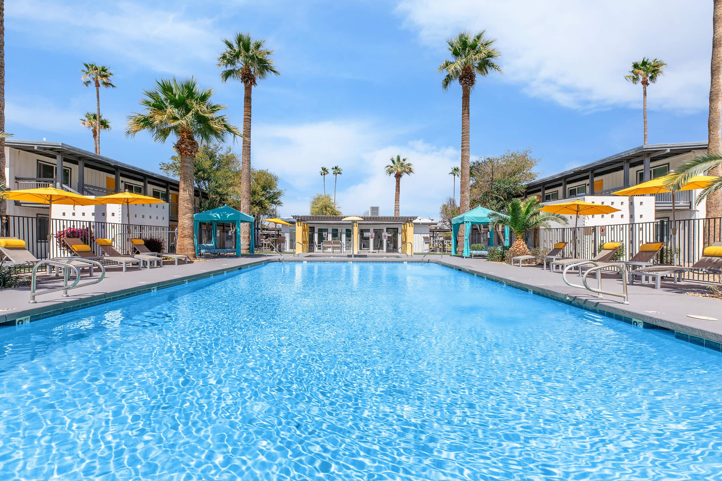 A serene swimming pool surrounded by palm trees and lounge chairs. Colorful umbrellas provide shade, and buildings are visible in the background under a clear blue sky. The scene evokes a relaxed and inviting atmosphere.