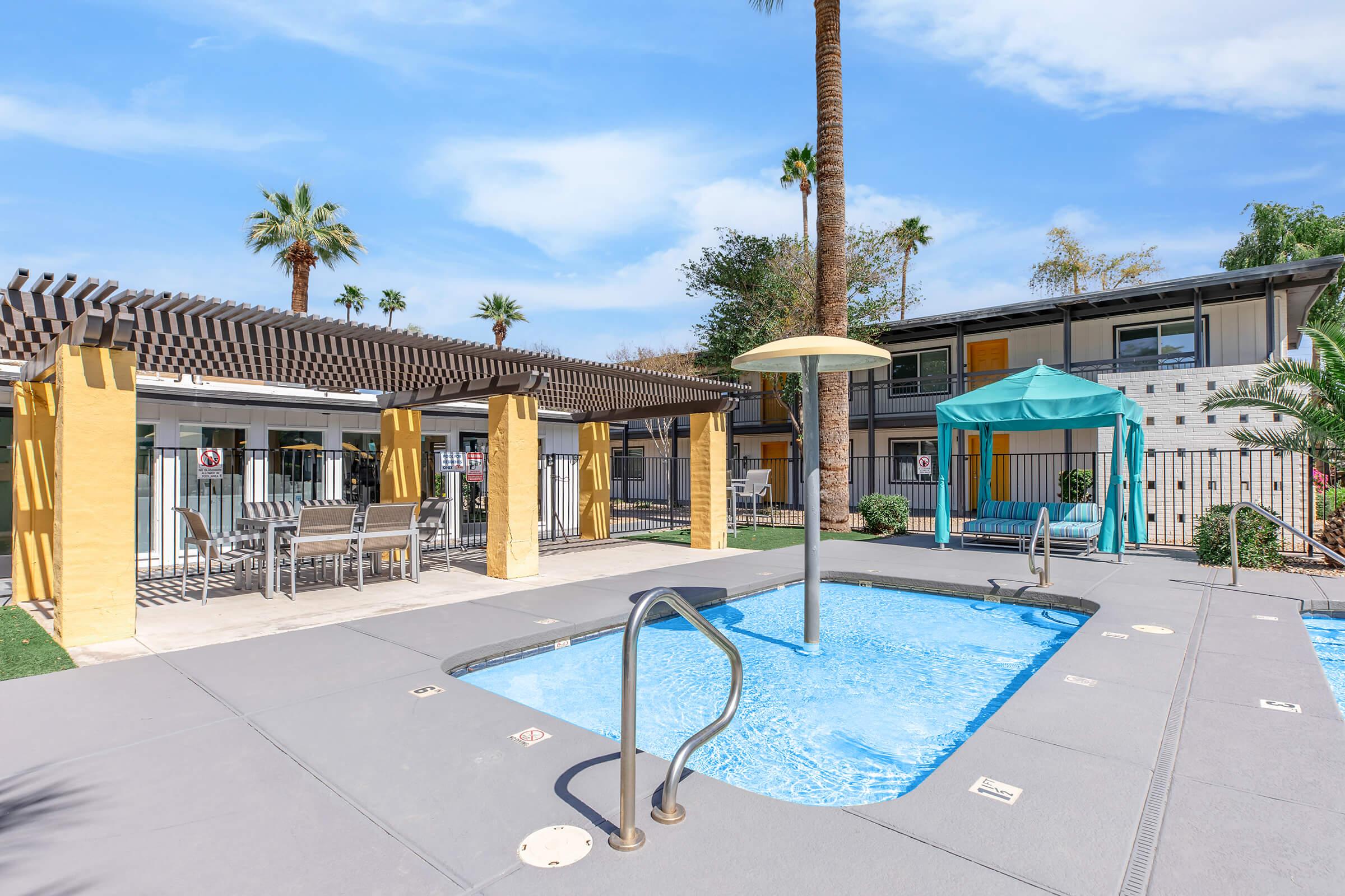 A clear blue pool area with a shallow wading section, shaded by a pergola and surrounded by palm trees. Nearby, there’s a blue cabana and patio furniture for relaxation. The setting is sunny, giving a vibrant and inviting atmosphere.