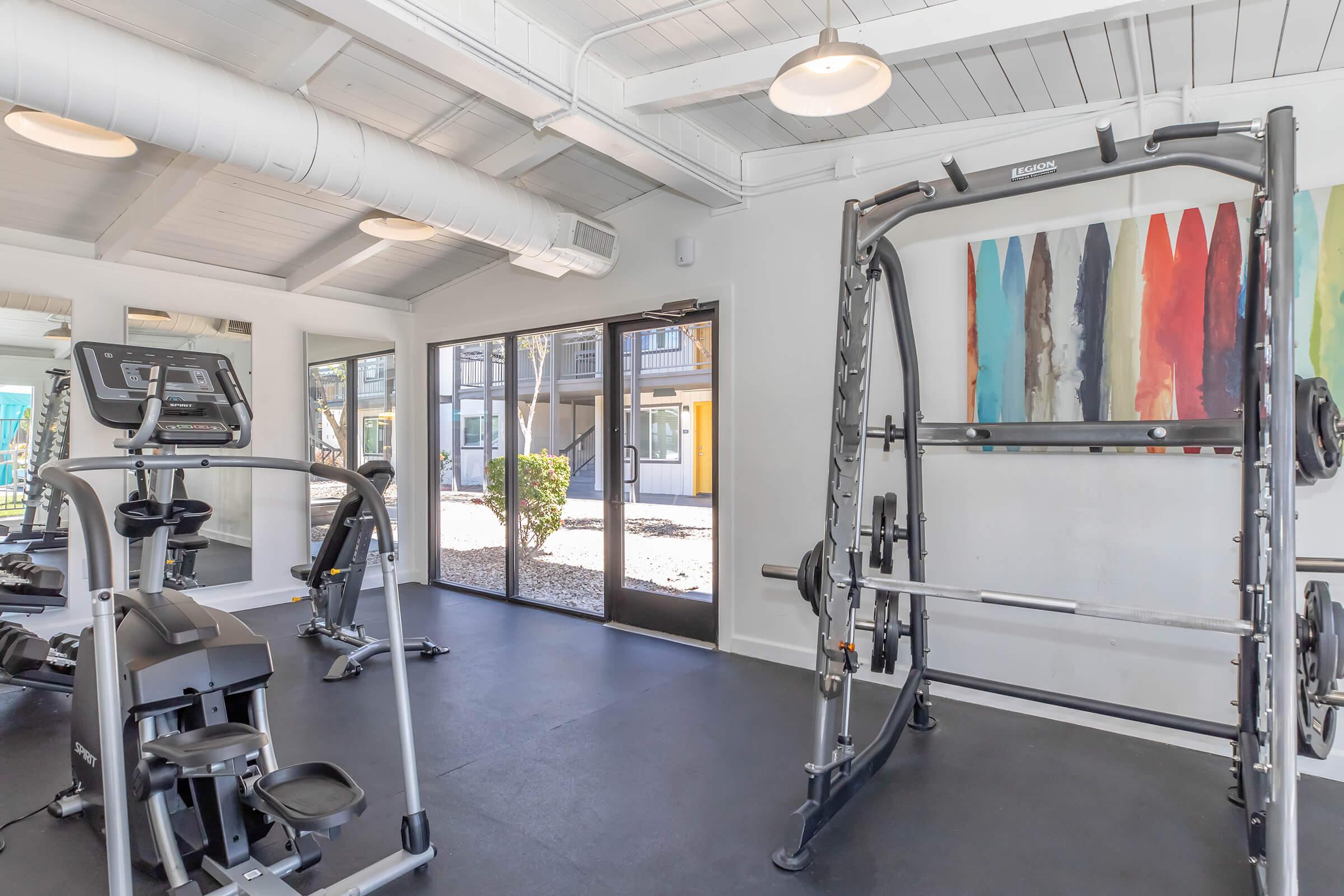 A modern gym interior featuring various workout machines, including a treadmill and weight training equipment. Large windows provide natural light, with a view of the outdoors. The wall is adorned with colorful abstract art, enhancing the energetic atmosphere of the space.