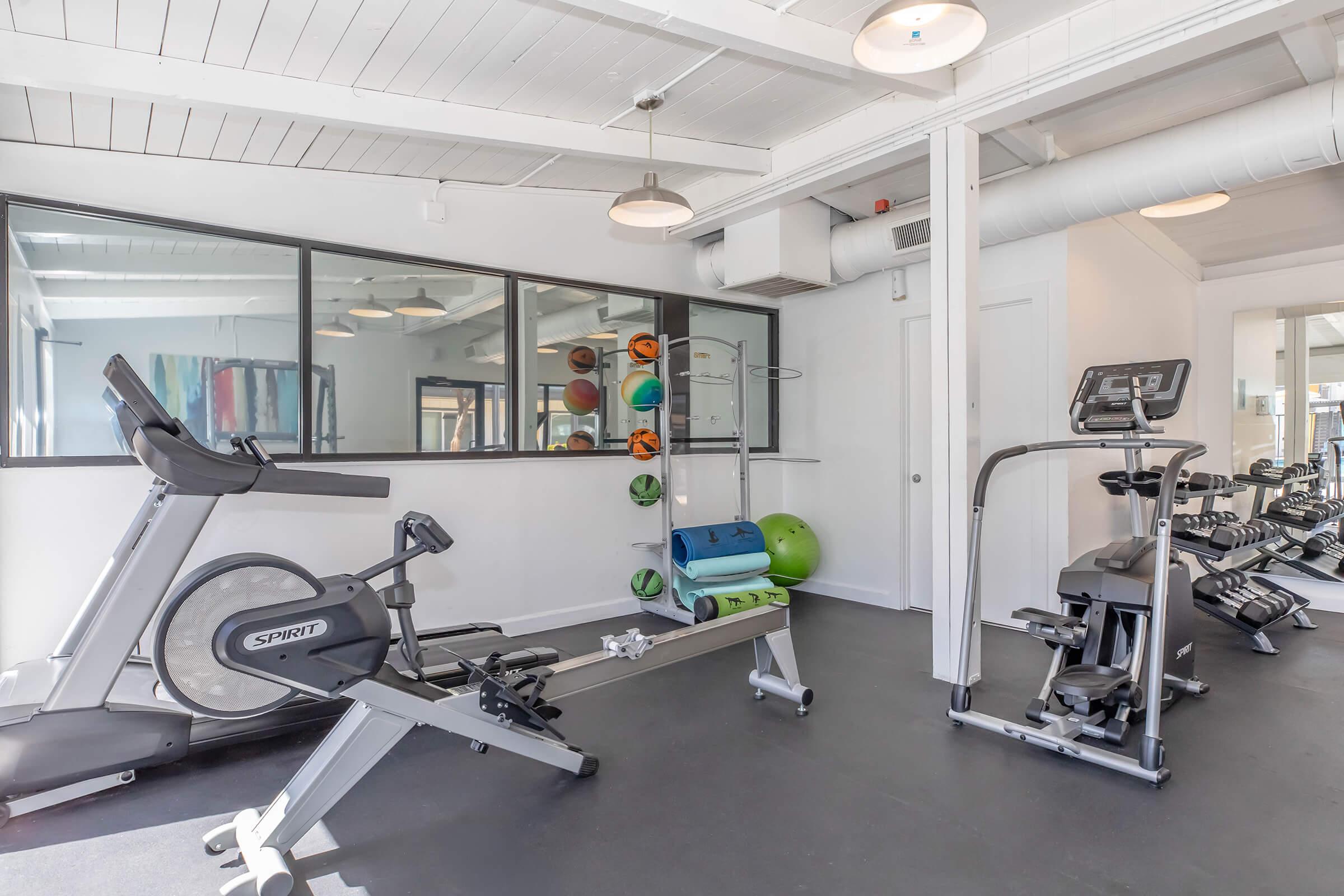 A modern gym interior featuring exercise equipment including an elliptical machine, weight bench, and a treadmill. The space has large windows and is well-lit, with fitness accessories like weights and a stability ball visible. The floor is black, creating a sleek and contemporary atmosphere.