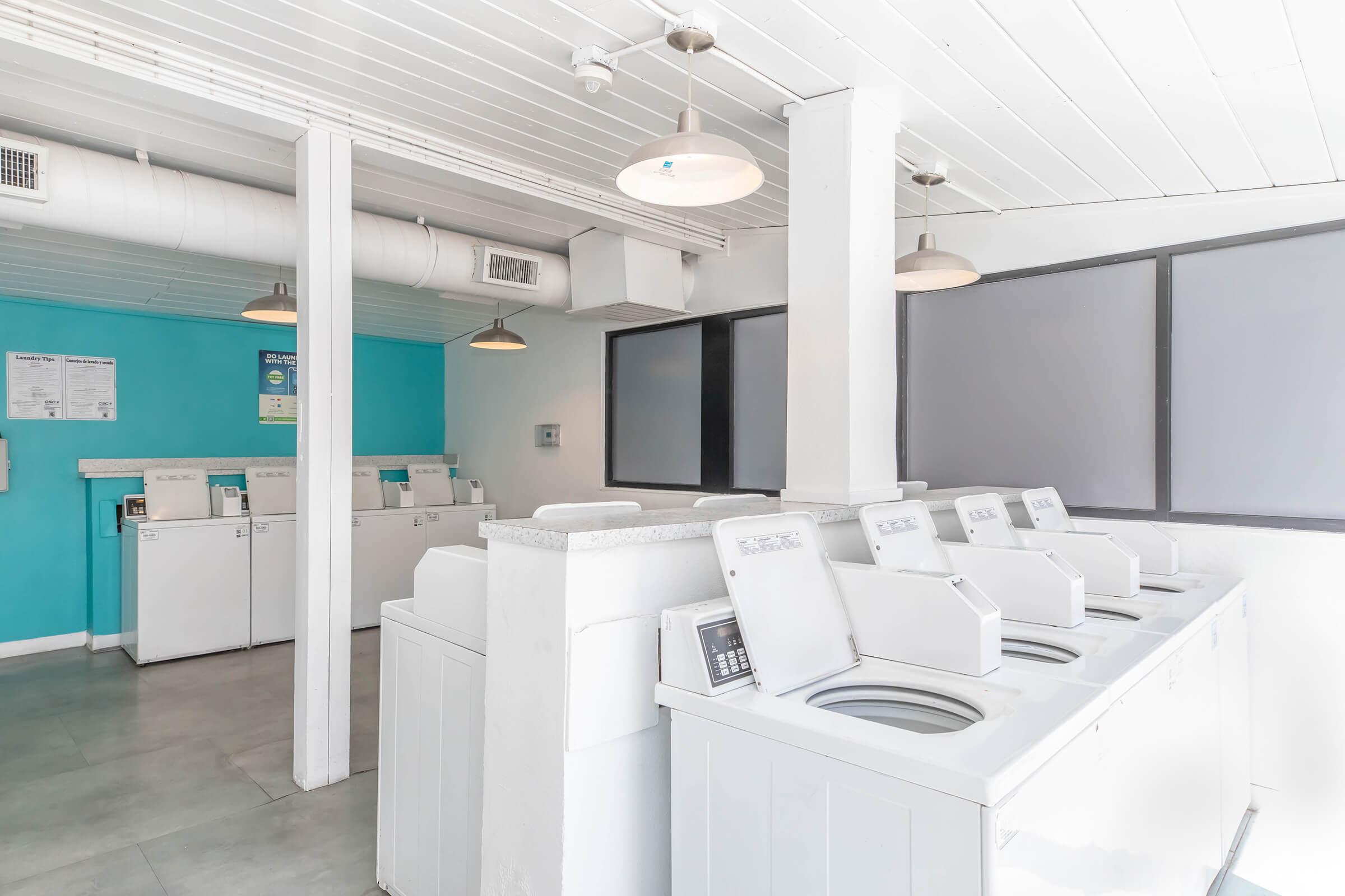 A clean, well-lit laundry room featuring several white washing machines lined up against a wall. The backdrop has a light blue paint, and overhead lights provide bright illumination. Visible on the walls are notices or instructions related to laundry usage. The room has a modern, minimalist aesthetic.