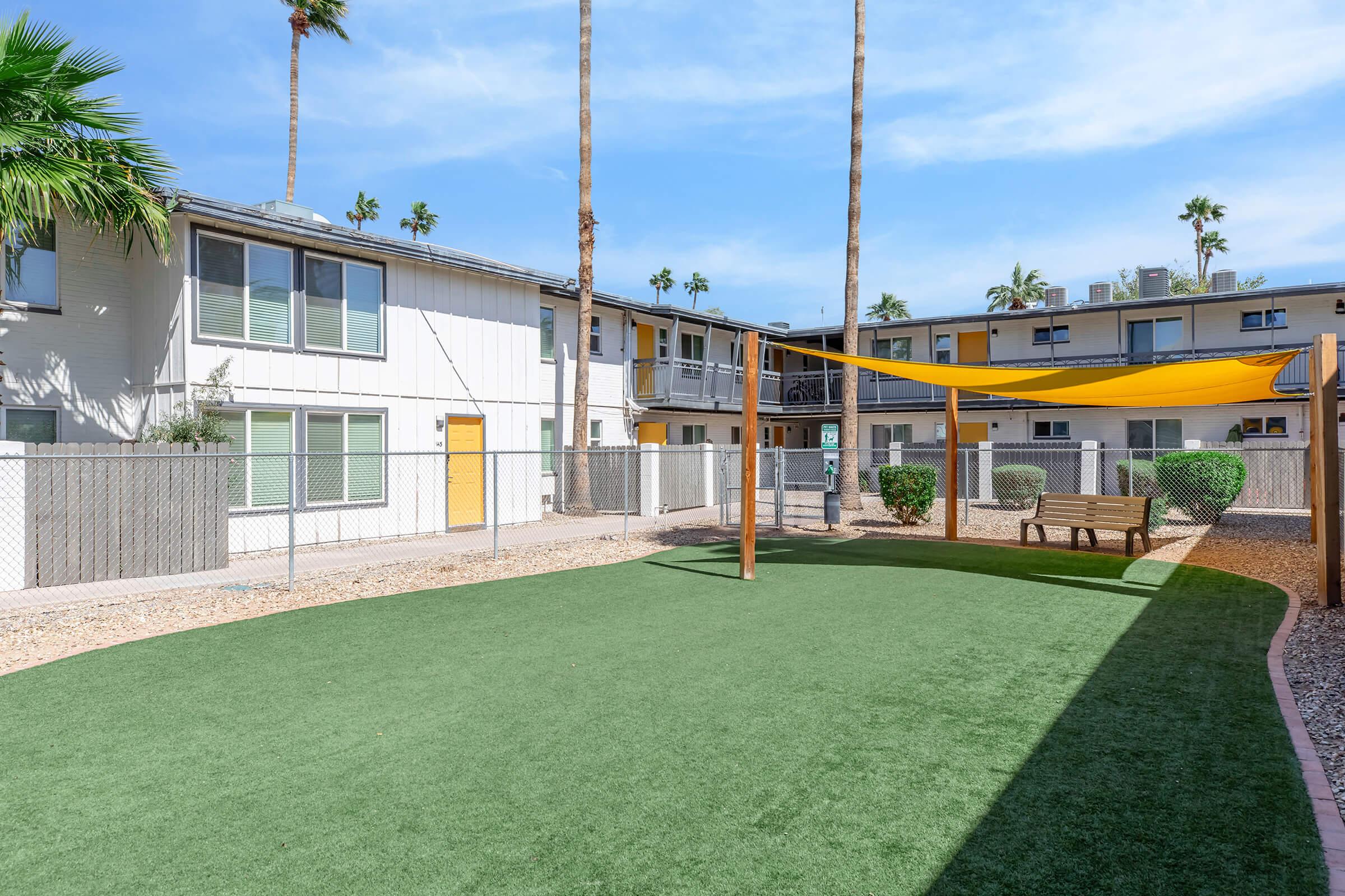 A spacious outdoor area featuring green artificial turf, surrounded by low fences and palm trees. In the foreground, there's a wooden swing set and a bench under a shaded area. The background shows two-story apartment buildings with light-colored siding and balconies.