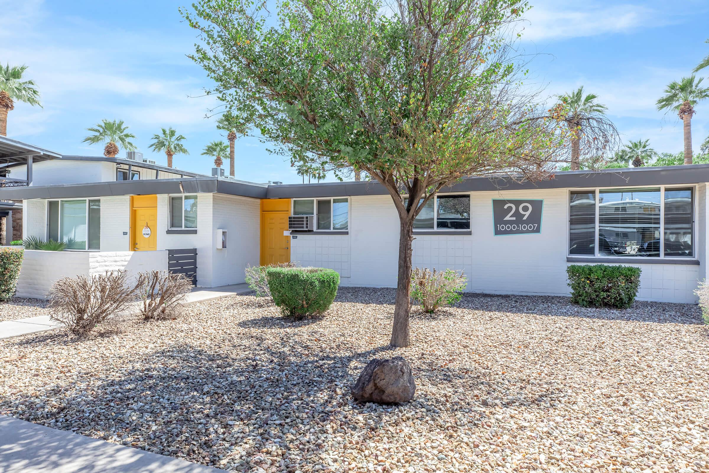 Exterior view of a mid-century modern style house featuring a gravel landscape. The home has a yellow and gray facade, with the house number "29" displayed prominently. A small tree and shrubs complement the entrance, and palm trees are visible in the background, indicating a sunny climate.