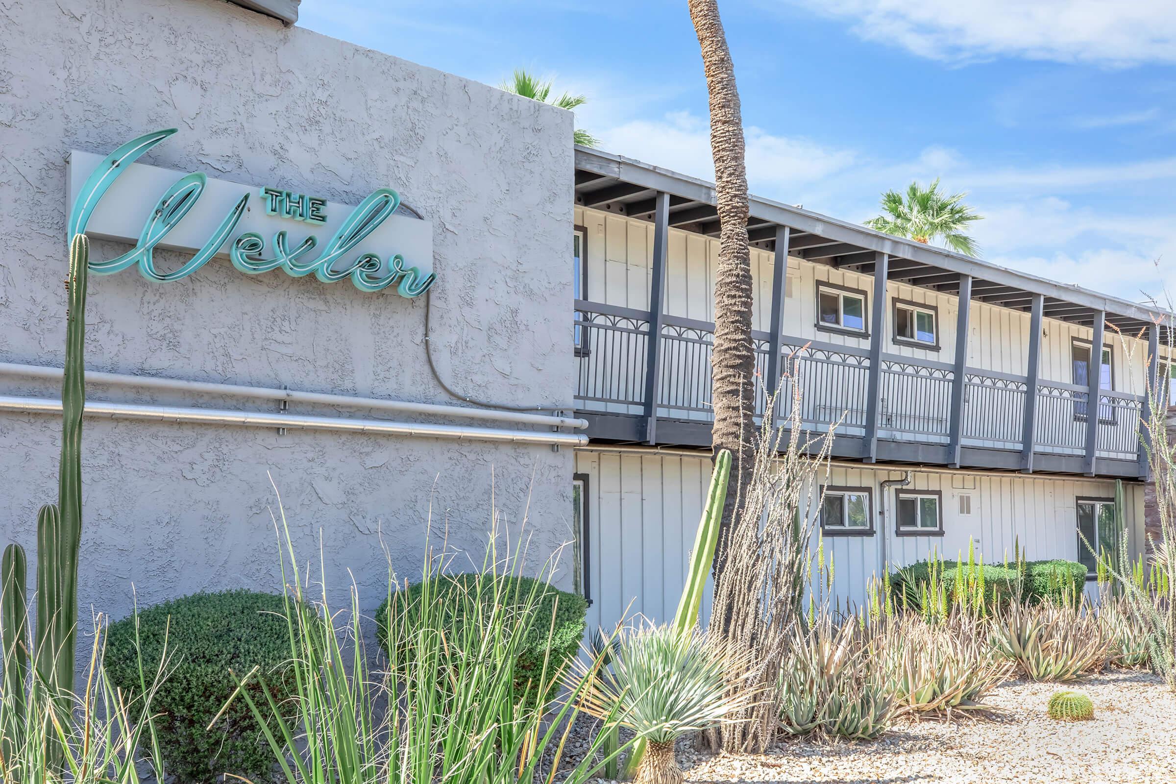 The image features the exterior of a building with a sign that says "The Wexler." The structure has a modern design with a balcony and is surrounded by desert landscaping, including cacti and other plants. Palm trees are visible in the background, under a clear blue sky.