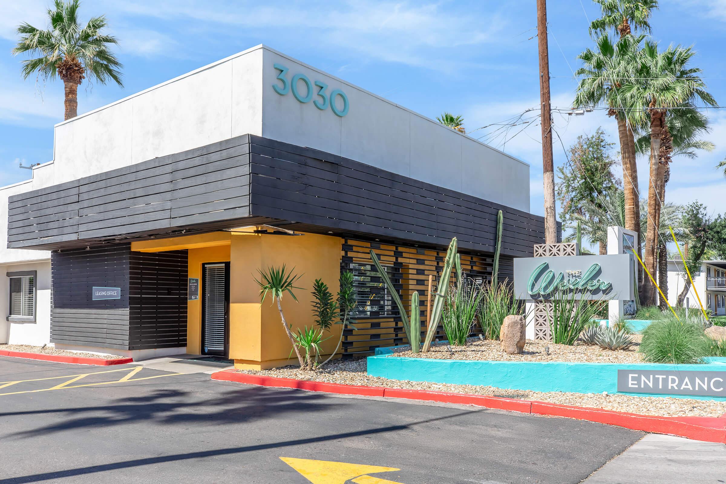 Modern building exterior with a minimalist design, featuring a vibrant yellow entrance, large windows, and a landscaped area with desert plants. The address "3030" is displayed prominently above the entrance, and there are palm trees and other greenery around the property, adding a tropical feel.