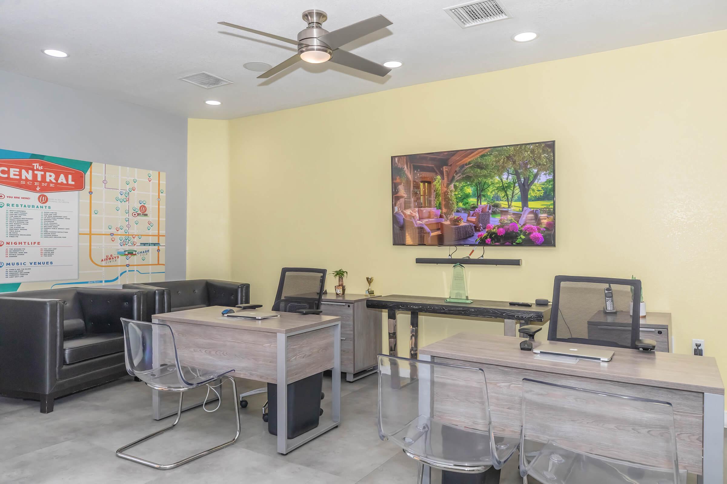 Modern office space featuring two clear acrylic desks with chairs, a large flat-screen TV on the wall displaying a scenic outdoor view, a potted plant, and a cozy seating area with black leather couches. A wall map titled "The Central" is visible, providing information about local amenities.