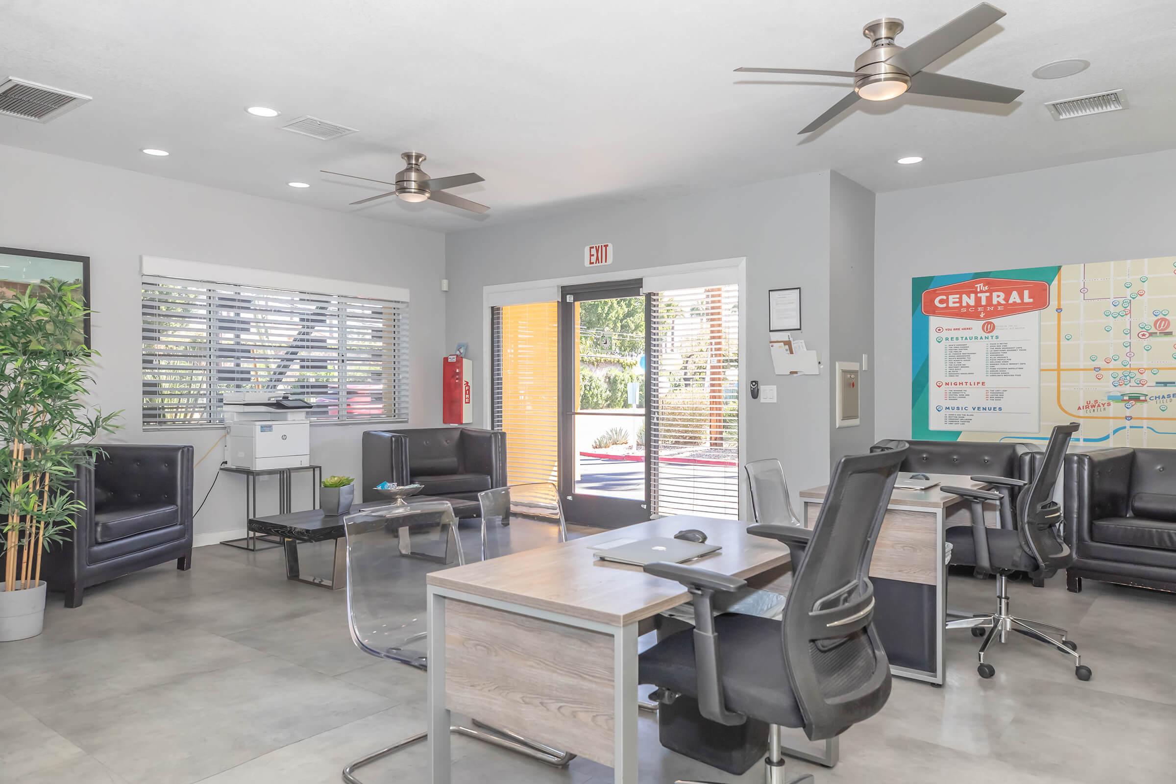 Modern office interior featuring two black leather chairs, glass-top desks with black office chairs, and a printer. The room is bright with large windows and plants, casting natural light. A door leads to an outdoor area, and a wall map labeled "The Central" is visible. Ceiling fans are installed for ventilation.