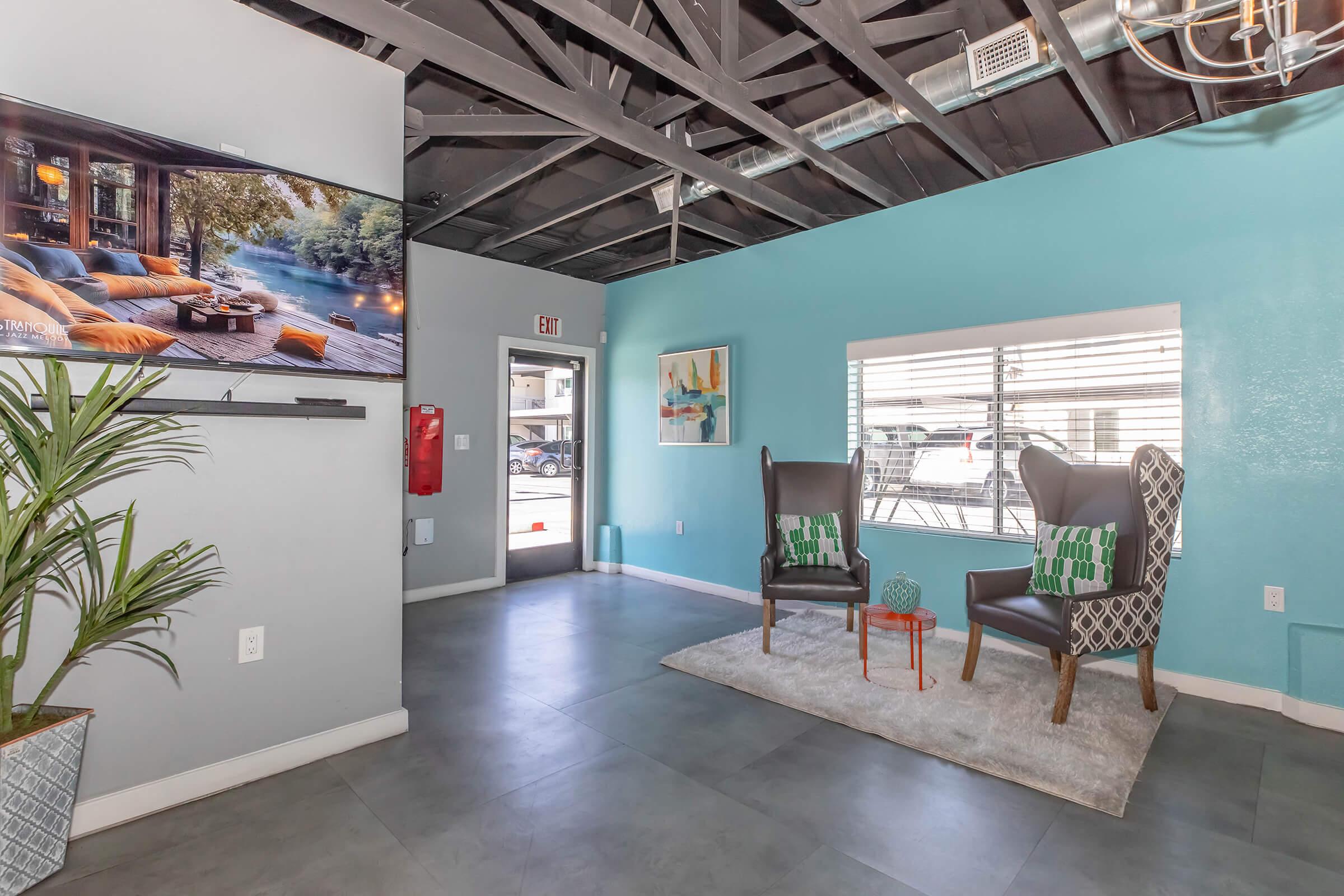 A modern lobby area featuring two stylish armchairs with patterned cushions, a small orange table between them, and a large wall-mounted television displaying an outdoor scene. The walls are painted in a calming turquoise, and there are potted plants, adding a touch of greenery to the space.