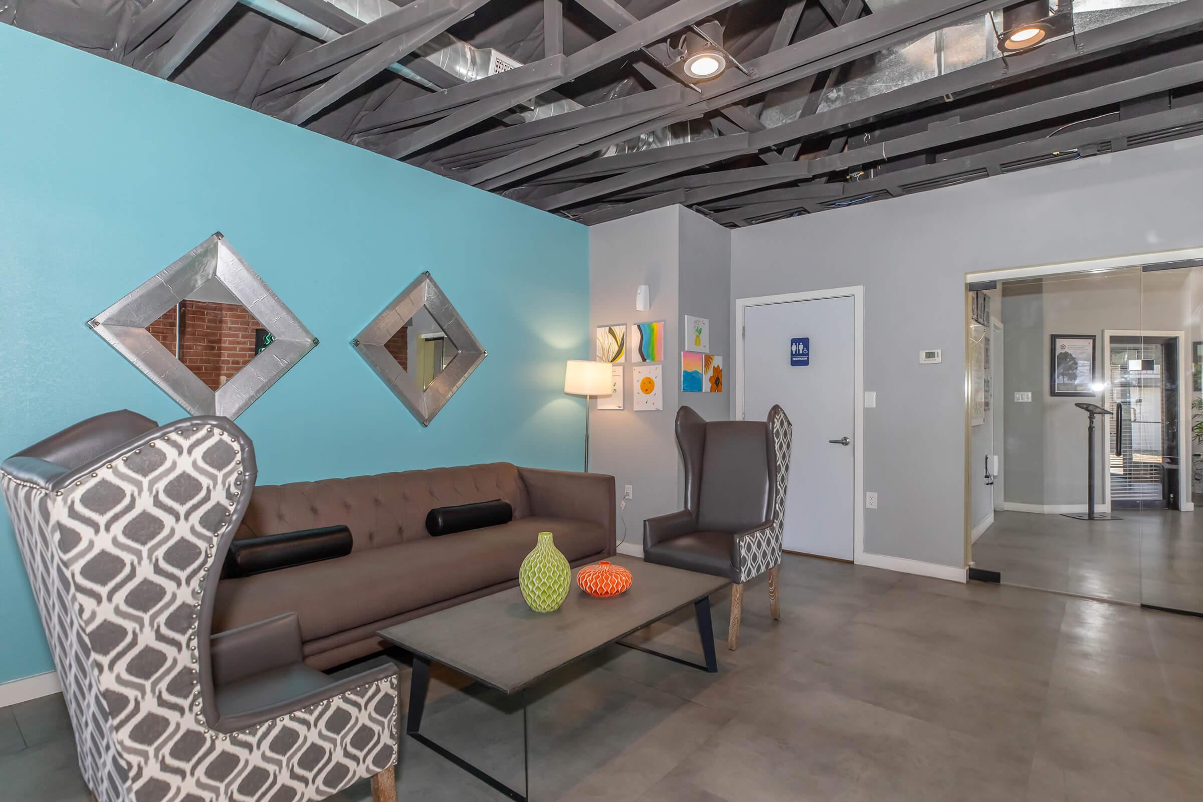 A modern lounge area featuring a gray patterned couch with black cushions, a glass coffee table with colorful decor items, and two diamond-shaped mirrors on the turquoise wall. The space has exposed beams in the ceiling and leads to a doorway with a sign, indicating the presence of additional rooms.