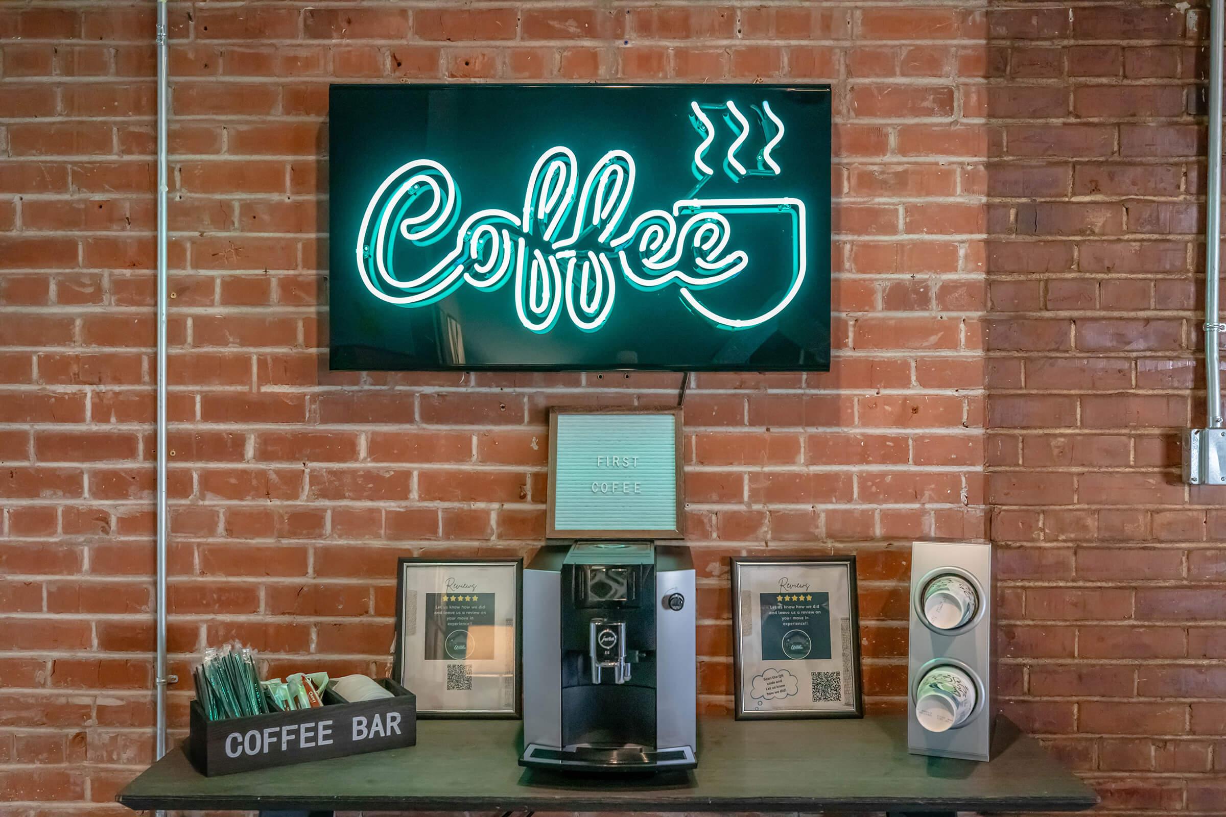 Neon sign that says "Coffee" with steam above a coffee cup, displayed on a brick wall. Below, a coffee machine sits on a table with a "Coffee Bar" sign, framed artwork, and stacking cups organized on the side. The atmosphere is cozy and inviting, perfect for coffee enthusiasts.