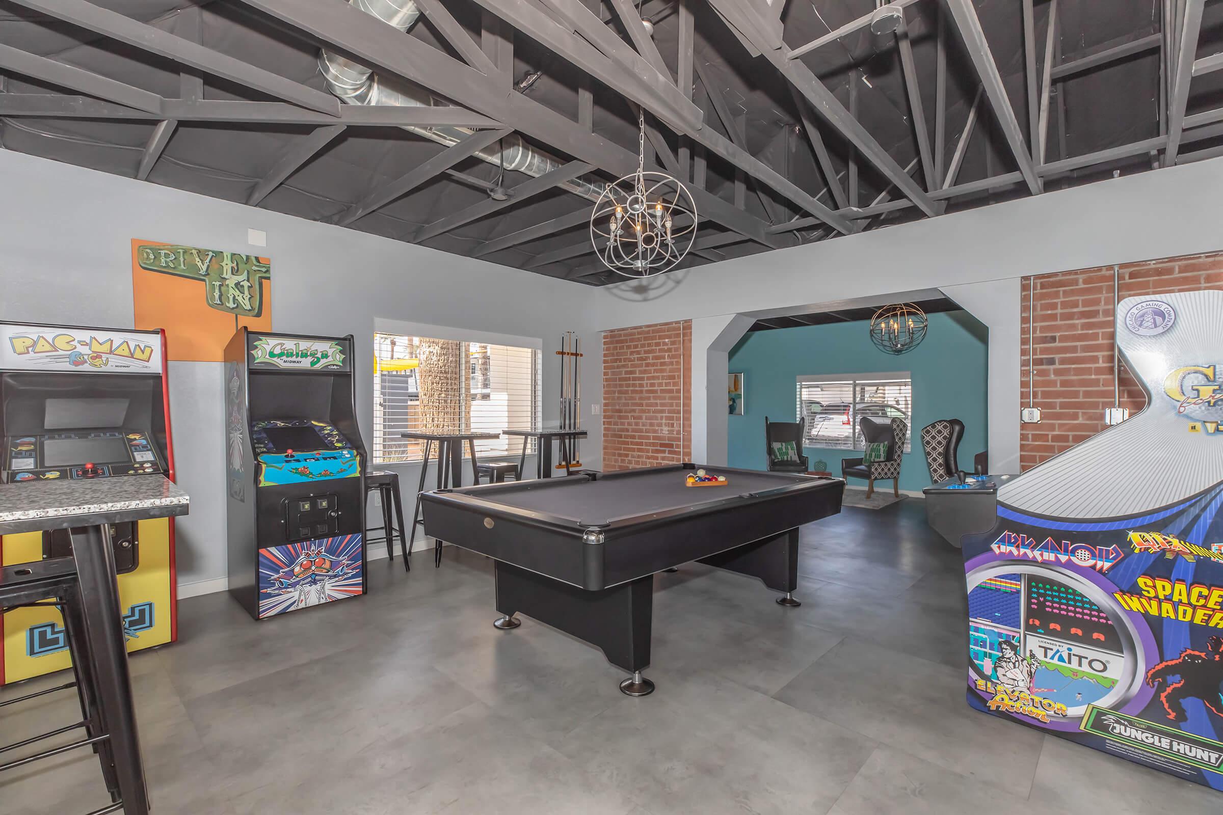 A lively game room featuring a black pool table at the center, vintage arcade machines including Pac-Man and Space Invaders, exposed brick walls, and modern light fixtures. Comfortable seating is visible in the background, creating a fun and inviting recreational space.
