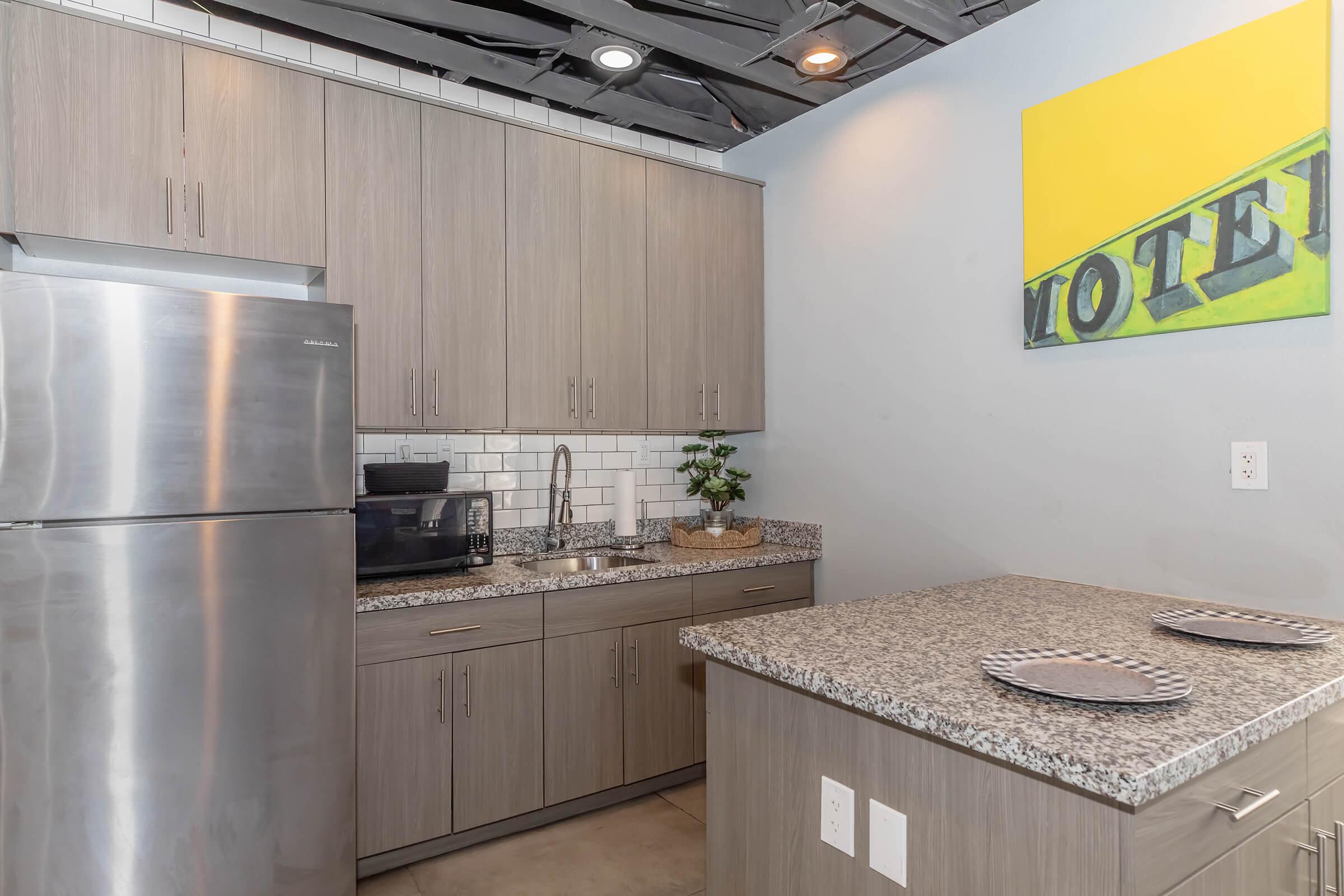 Modern kitchen featuring stainless steel appliances, including a refrigerator and coffee maker. The countertop is made of gray speckled granite, with two plates set on it. There's a small plant and artwork on the wall that reads "VOTE." The cabinetry is light wood, and the backsplash is white subway tile.