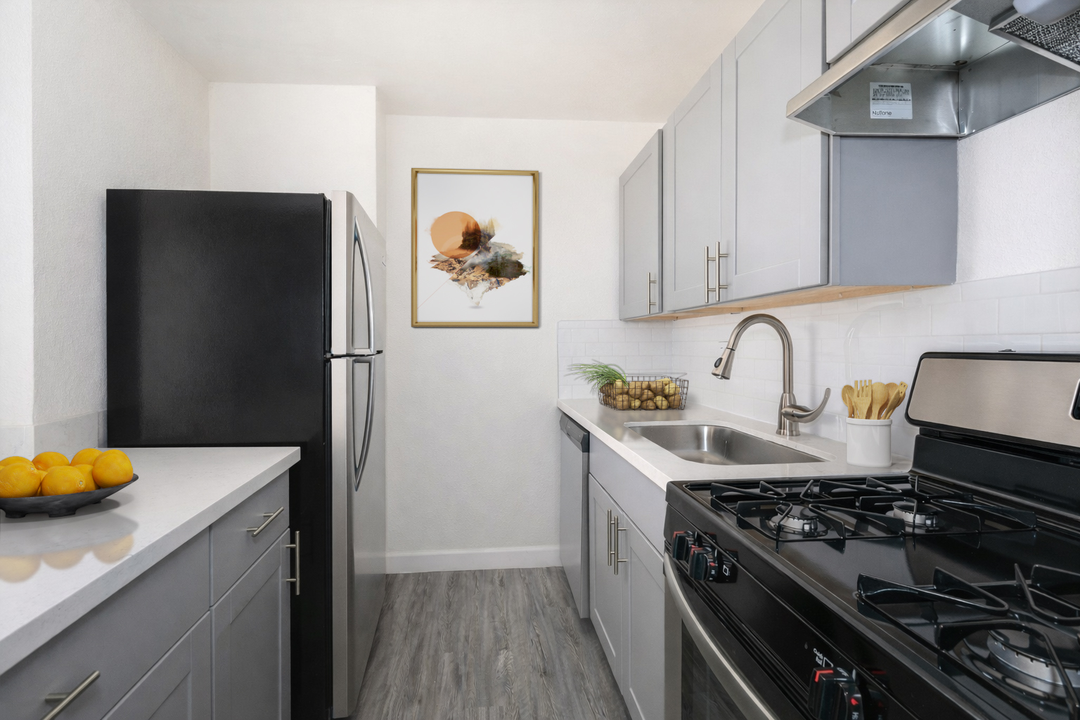 Modern kitchen featuring gray cabinets, stainless steel appliances, a white countertop, and a sink. A framed wall art piece hangs above the sink. A bowl of oranges is on the countertop, with hexagonal tiles and wood-like flooring completing the aesthetic.