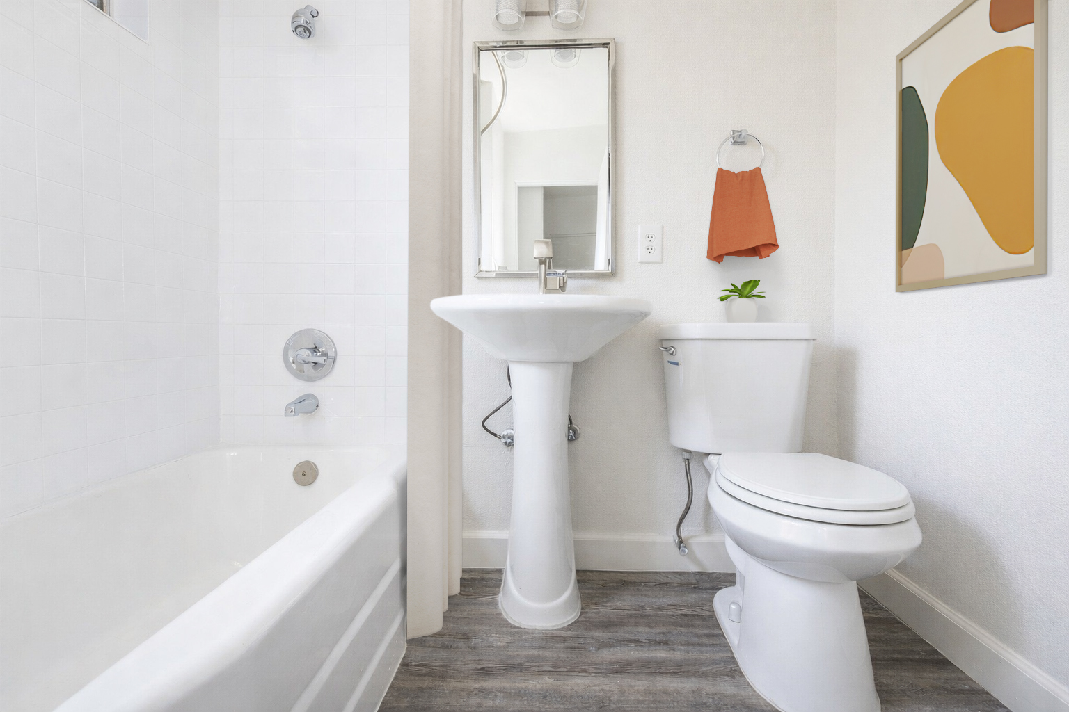 A modern bathroom featuring a bathtub, pedestal sink, and a toilet. The walls are light-colored, with a decorative wall art piece on the right. A small plant is placed on the sink, and there is a towel hanging on a rack beside the mirror. Natural light brightens the space.