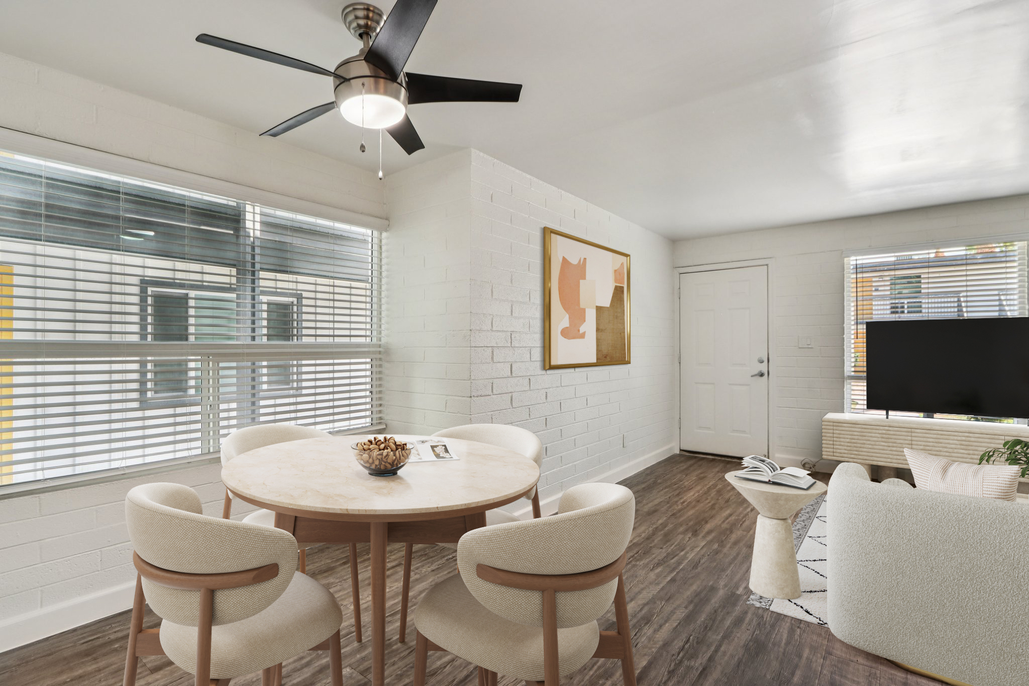 A modern dining area featuring a round table with a decorative centerpiece, surrounded by beige upholstered chairs. A large window with horizontal blinds allows natural light to enter. In the background, there's a minimalist living area with a TV and a neutral-toned armchair, complemented by a wall-mounted artwork.