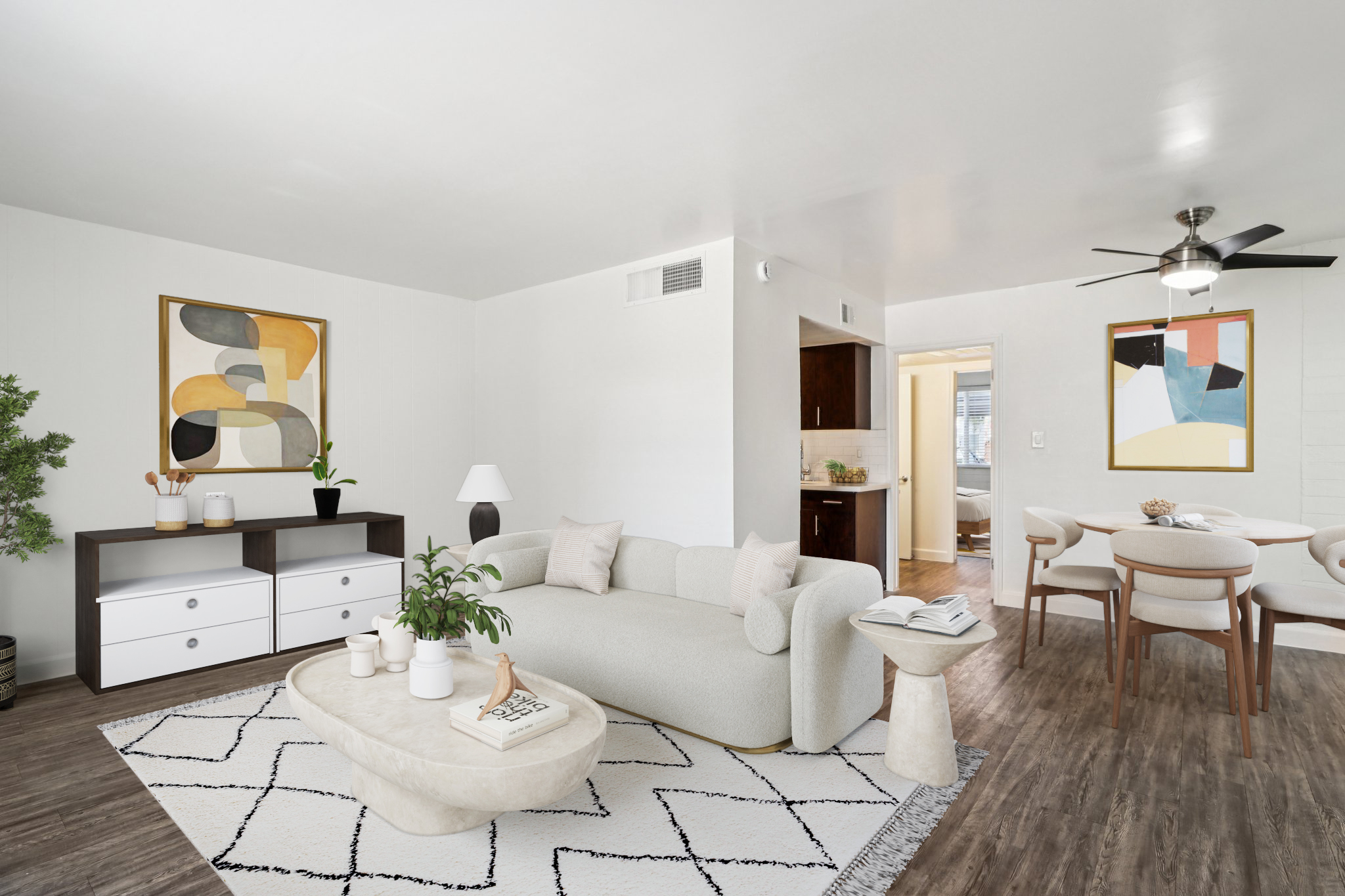A modern living room featuring a light-colored sofa, round coffee table, and a TV stand with drawers. On the walls, there are abstract art pieces. A plant adds greenery to the space, while a dining area with a round table and chairs is visible in the background. The room has stylish decor and a bright, inviting atmosphere.