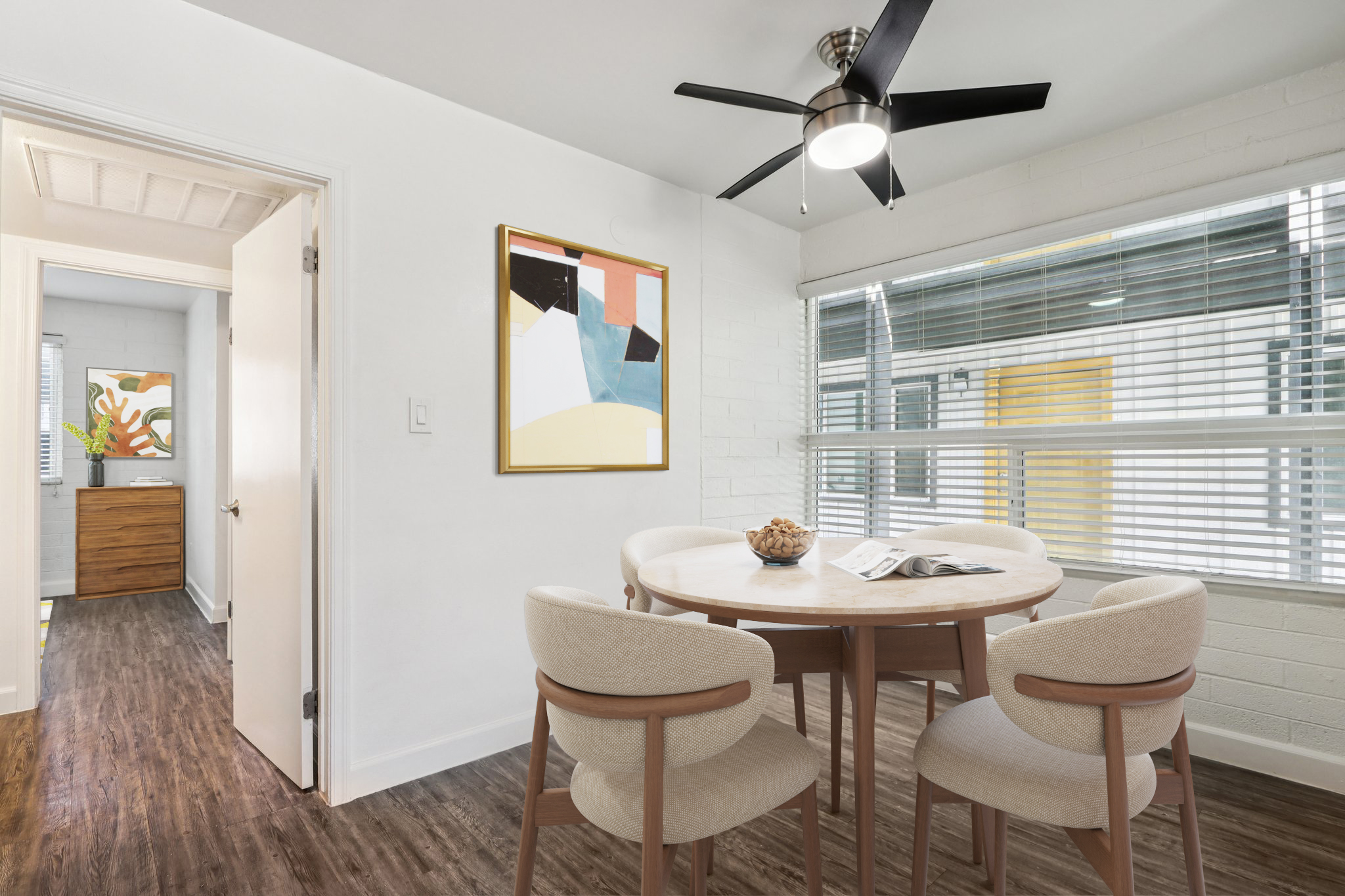 A modern dining area featuring a round wooden table surrounded by beige chairs. Large windows provide natural light, with a ceiling fan above. A framed abstract artwork hangs on the wall, and a doorway leads to another room with a stylish wooden dresser and colorful art. The floor is wooden, creating a warm ambiance.