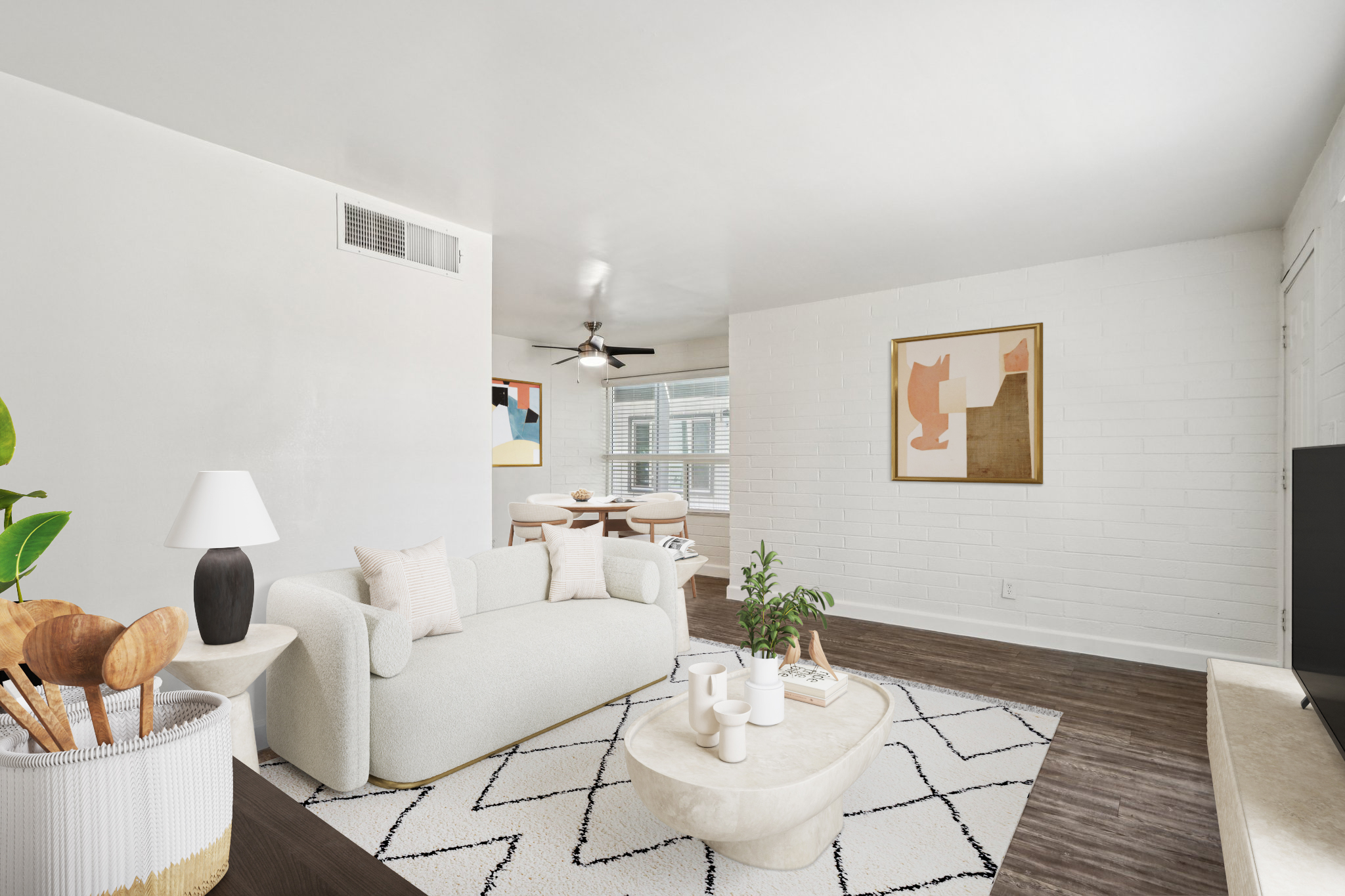 A modern living room with a light-colored couch, a round coffee table, and a decorative rug. There is a plant on the floor and a lamp on a side table. The room features large windows with natural light, a ceiling fan, and artwork on the wall. A dining area is visible in the background.