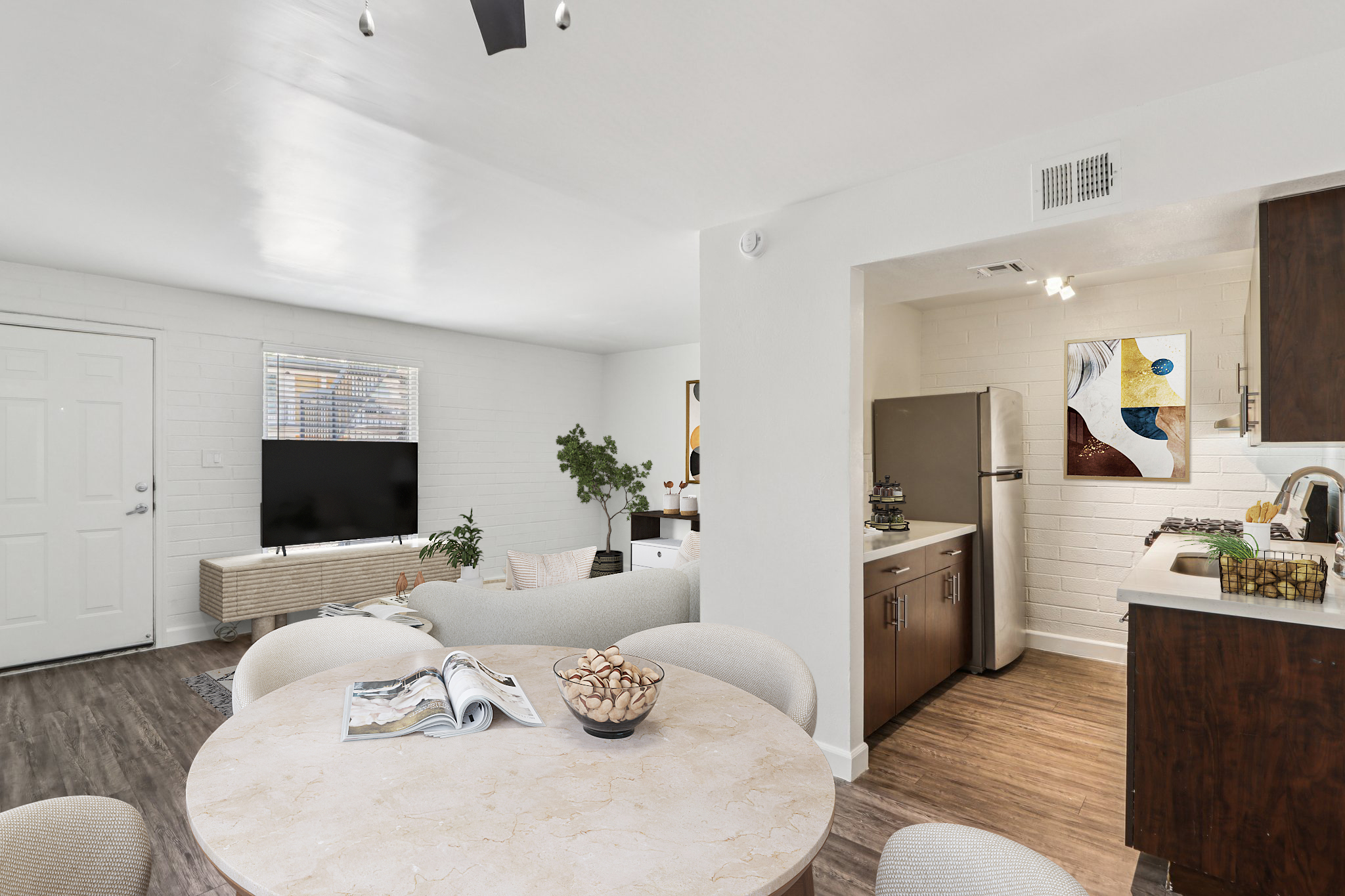 A modern, open-plan living space featuring a cozy seating area with a television, a round dining table with decorative items, and a kitchen with stainless steel appliances. The room is bright with white walls, and there's a piece of artwork on the kitchen wall, adding a touch of color.