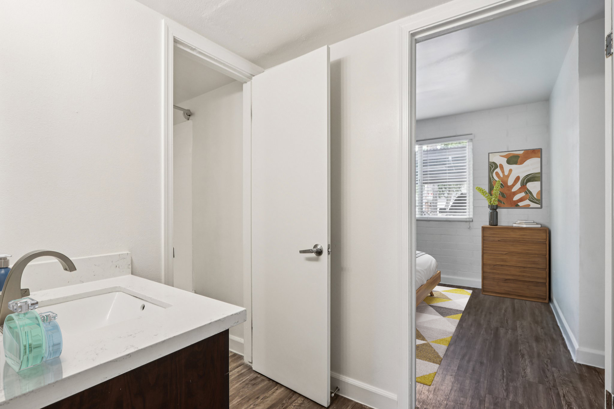 A bright, modern bathroom featuring a white sink area with a faucet and toiletries. A doorway leads into a simple, well-lit bedroom with a bed and a colorful artwork on the wall. The floors are wood-like, and natural light streams through a window in the bedroom.