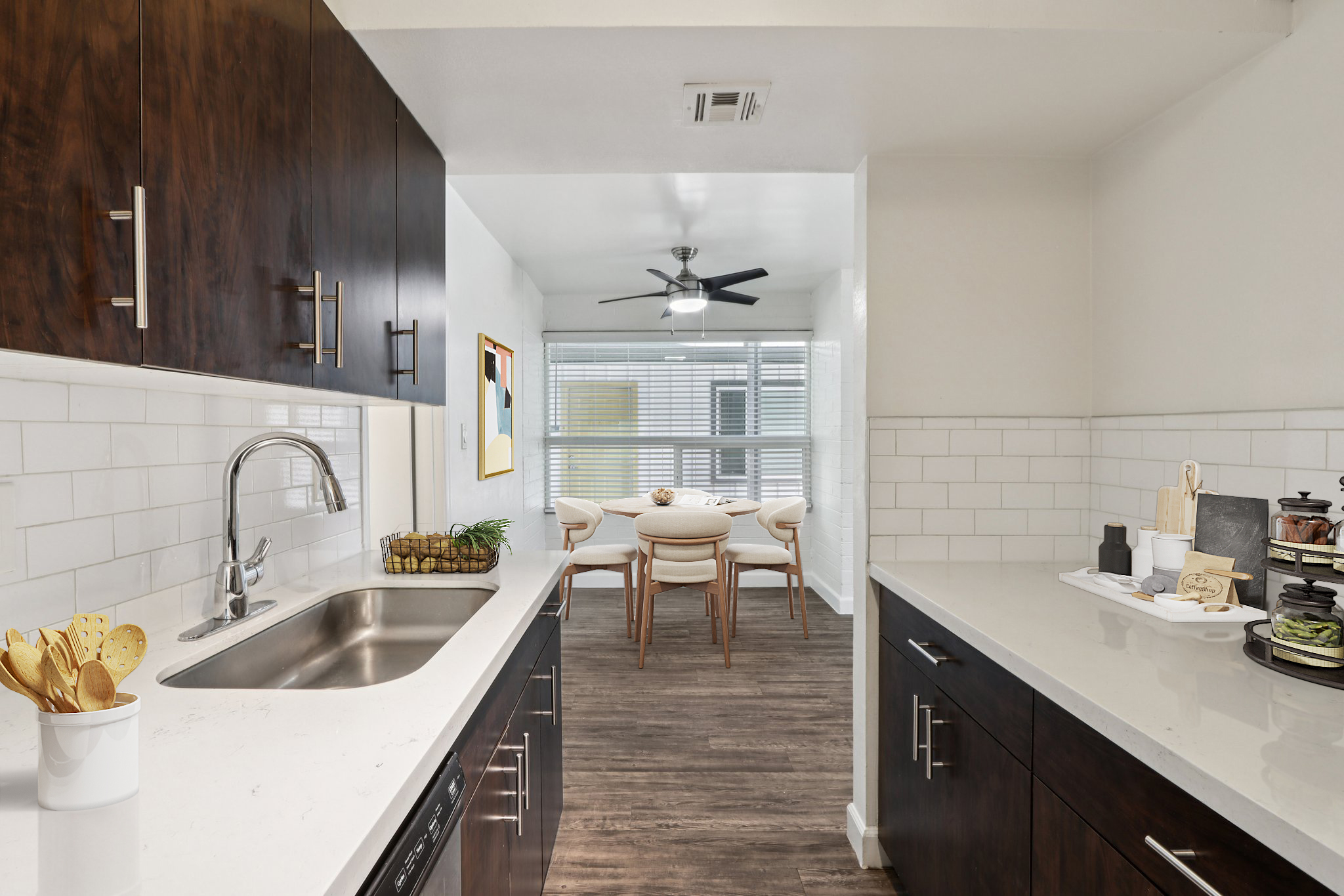 Modern kitchen with dark wood cabinets, white countertops, and a stainless steel sink. A small dining area is visible in the background featuring a round table and light-colored chairs, with a ceiling fan overhead and large windows providing natural light.