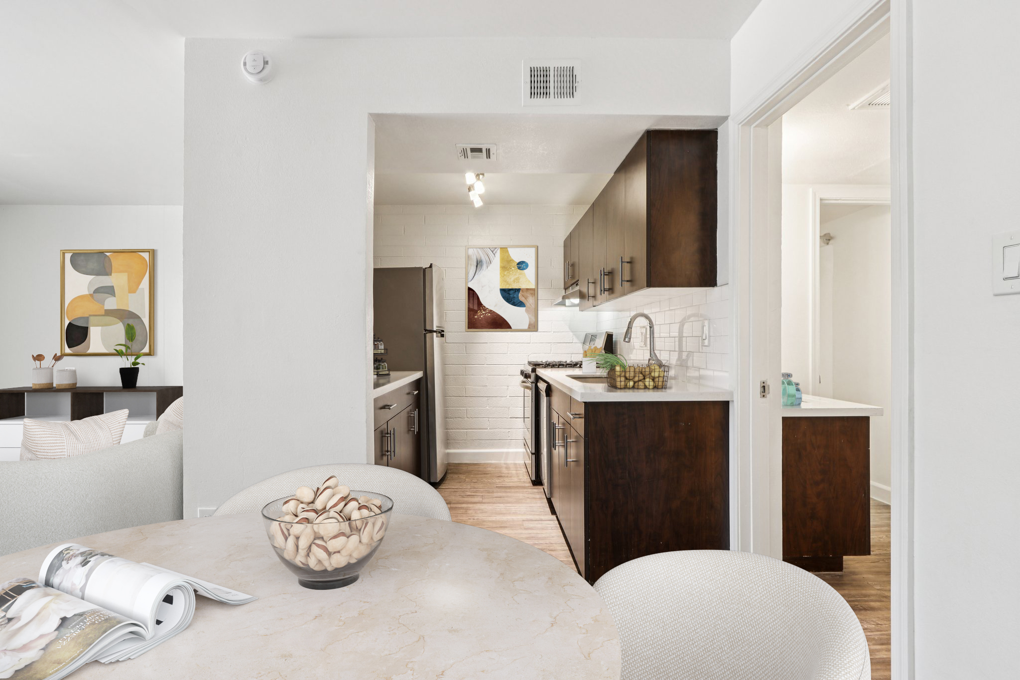 A modern kitchen area featuring dark wood cabinetry, stainless steel appliances, and white countertops. In the foreground, a round table with a decorative bowl of seashells sits in a dining area. The layout showcases an open space with artistic wall decor and a well-lit atmosphere.