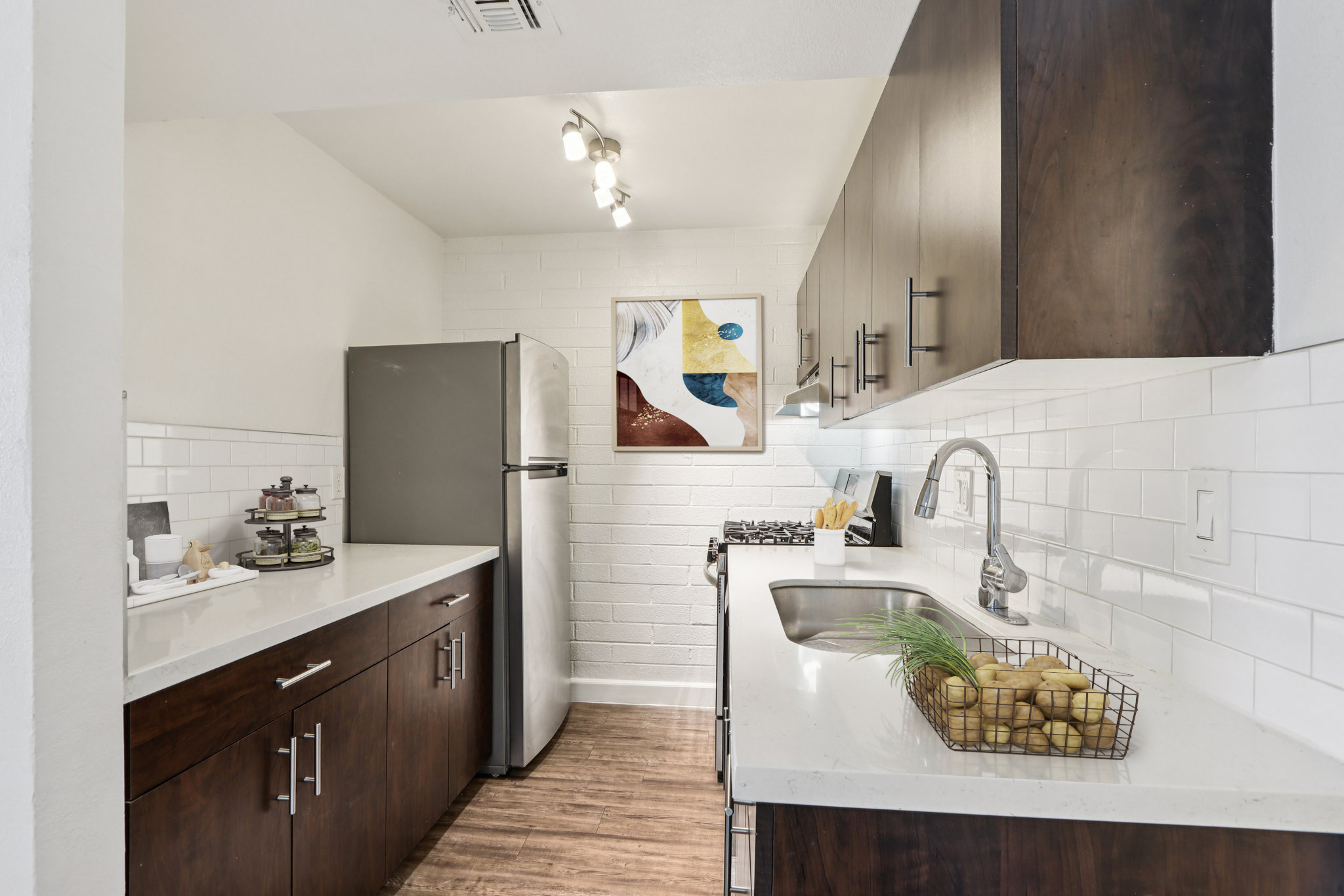 A modern kitchen with dark wooden cabinets, a stainless steel refrigerator, and a gas stove. The countertop features a basket of potatoes. A piece of abstract art hangs on the wall, complementing the bright, minimalist decor and white tiled backsplash. The space is well-lit, creating a warm and inviting atmosphere.