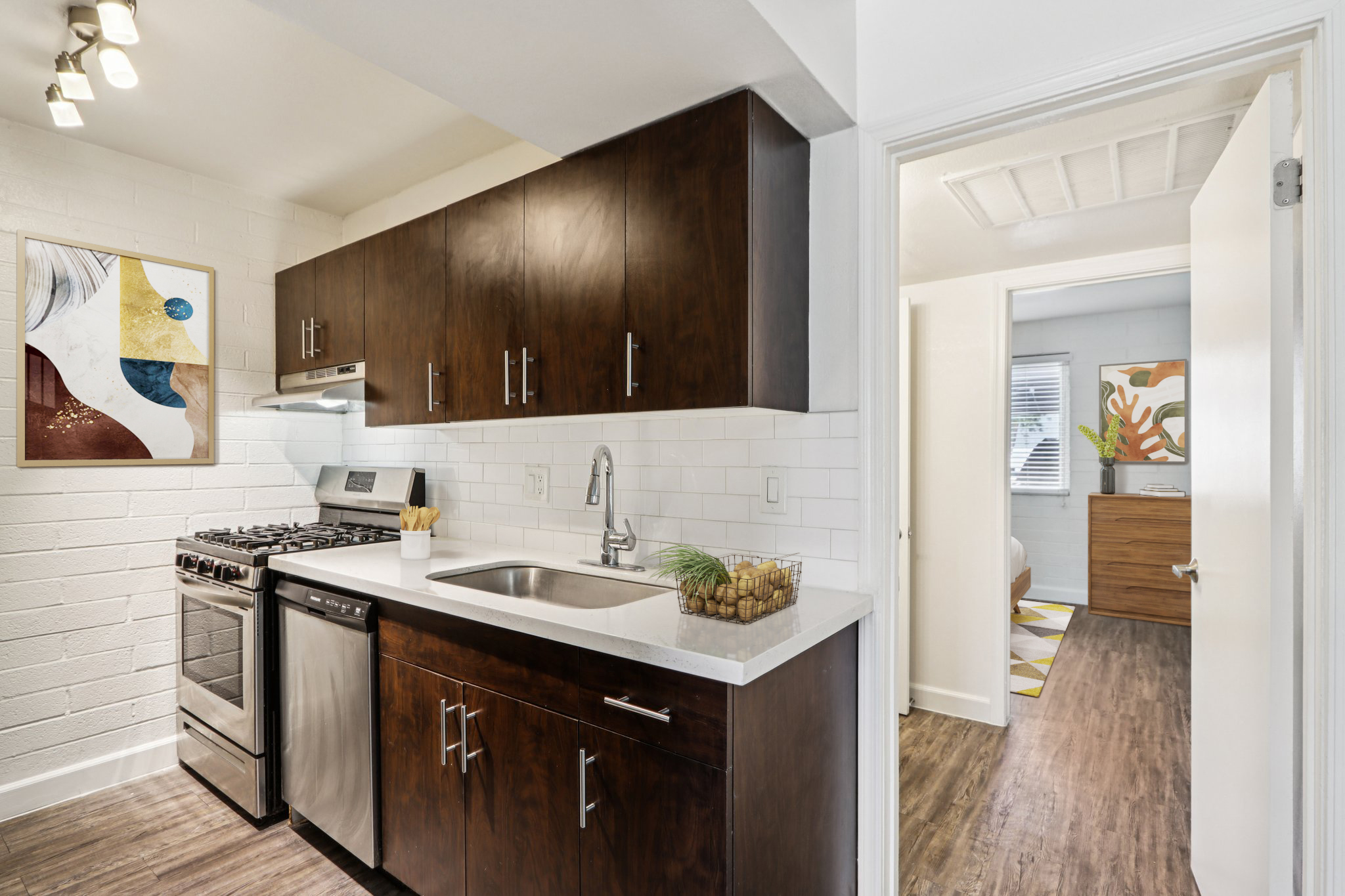 Modern kitchen with dark wood cabinets, stainless steel appliances, and a white countertop. Features a gas stove, sink, and decorative elements like a fruit basket. A doorway leads to another room with light-colored walls and art, glimpsed in the background.