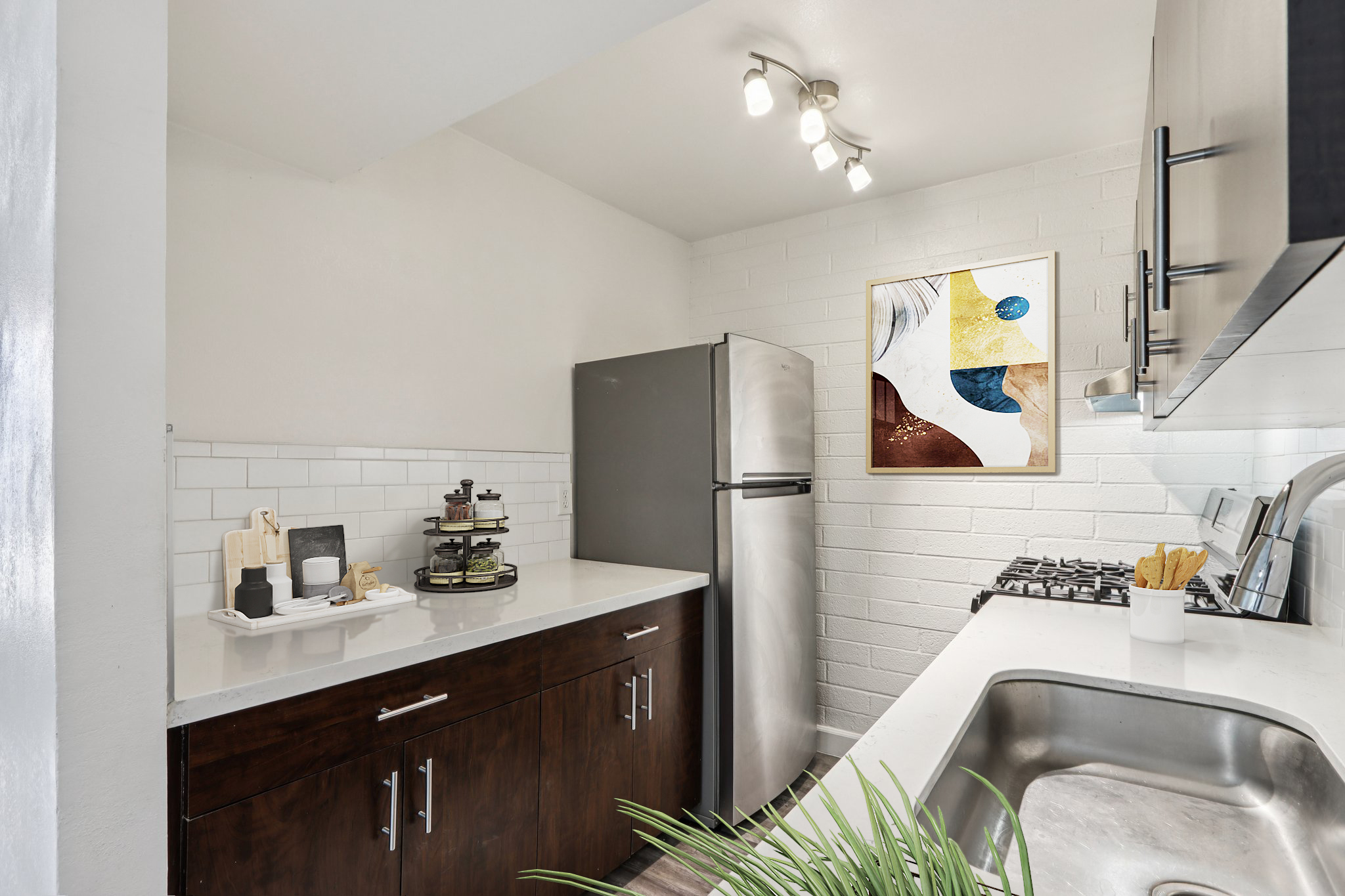 A modern kitchen featuring stainless steel appliances, including a refrigerator and gas stove. The countertops are light-colored, complemented by dark wooden cabinets. A decorative wall art piece is visible above the countertop, and a small plant adds a touch of greenery to the space.