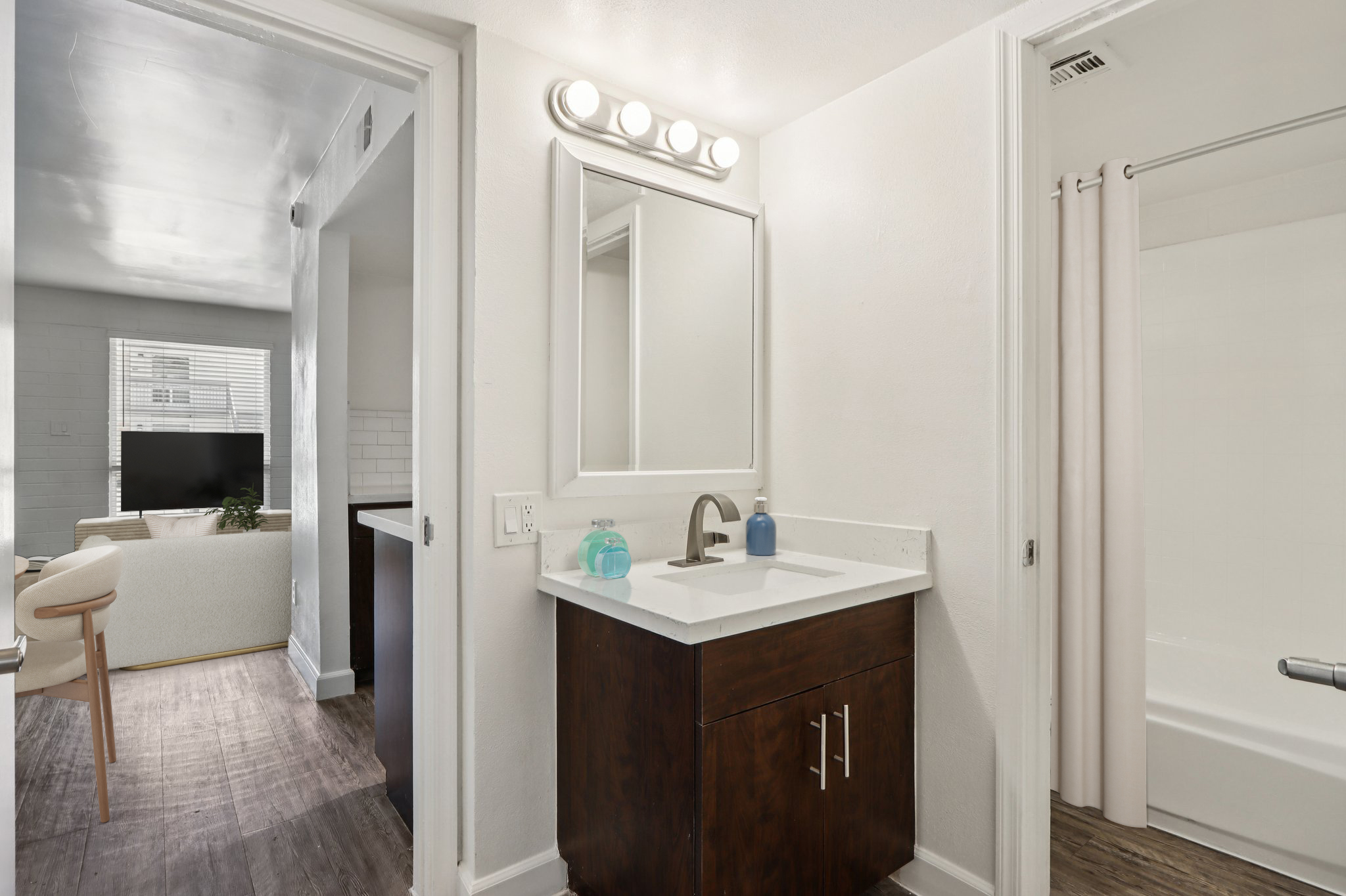 Interior view of a modern bathroom featuring a white sink with a dark wooden cabinet, a large mirror with overhead lighting, and a shower area. A doorway leads to a well-lit living space with a couch and a window visible in the background. The design has a clean, contemporary look with neutral colors.