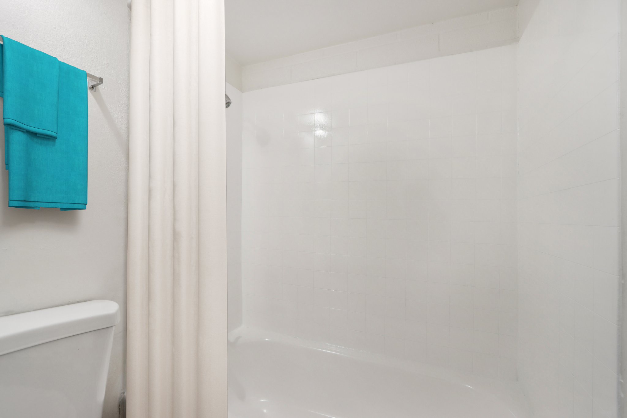 A clean and brightly lit bathroom with a shower area. The shower has a white curtain and textured white tiles. A blue towel is hanging on a rack nearby, adding a pop of color to the minimalist design of the space.