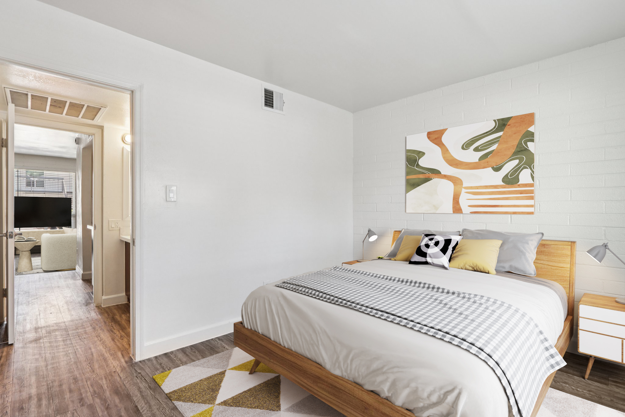 A bright and modern bedroom featuring a queen-sized bed with a light-colored comforter and decorative pillows. A colorful abstract artwork hangs above the bed. The room has light-colored walls and a wooden floor, with a bedside table and a lamp. A doorway leads to a bathroom area in the background.