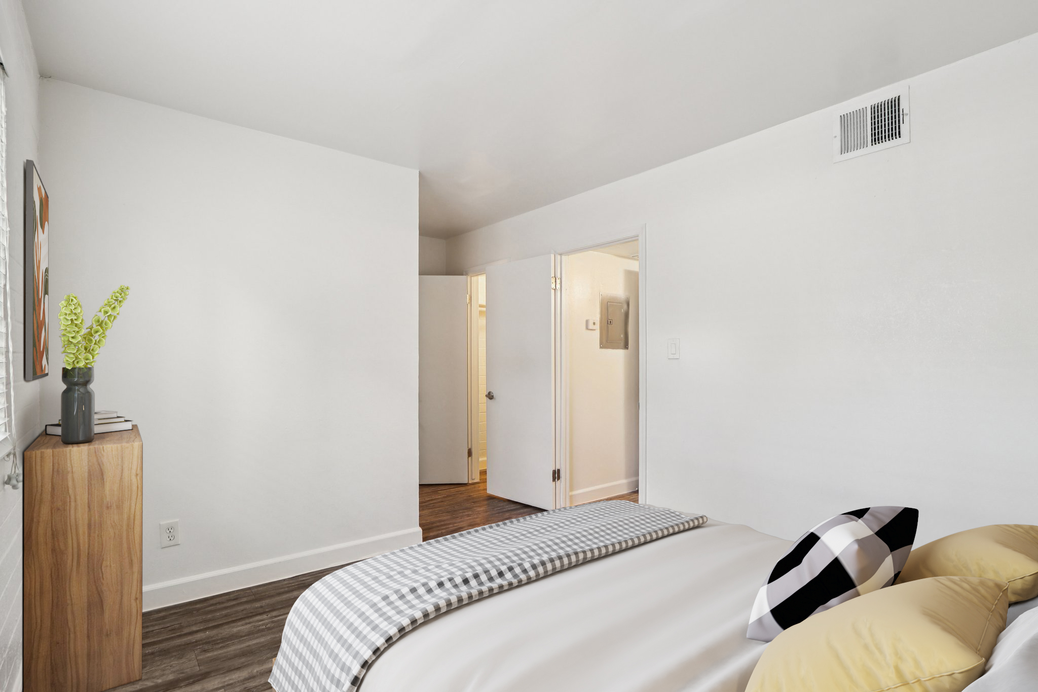 A modern bedroom featuring a neatly made bed with yellow and black accent pillows, a wooden bedside table with a plant, and a light-colored wall. A doorway in the background leads to another room, contributing to a bright and inviting atmosphere. The flooring is dark and enhances the room's contemporary feel.
