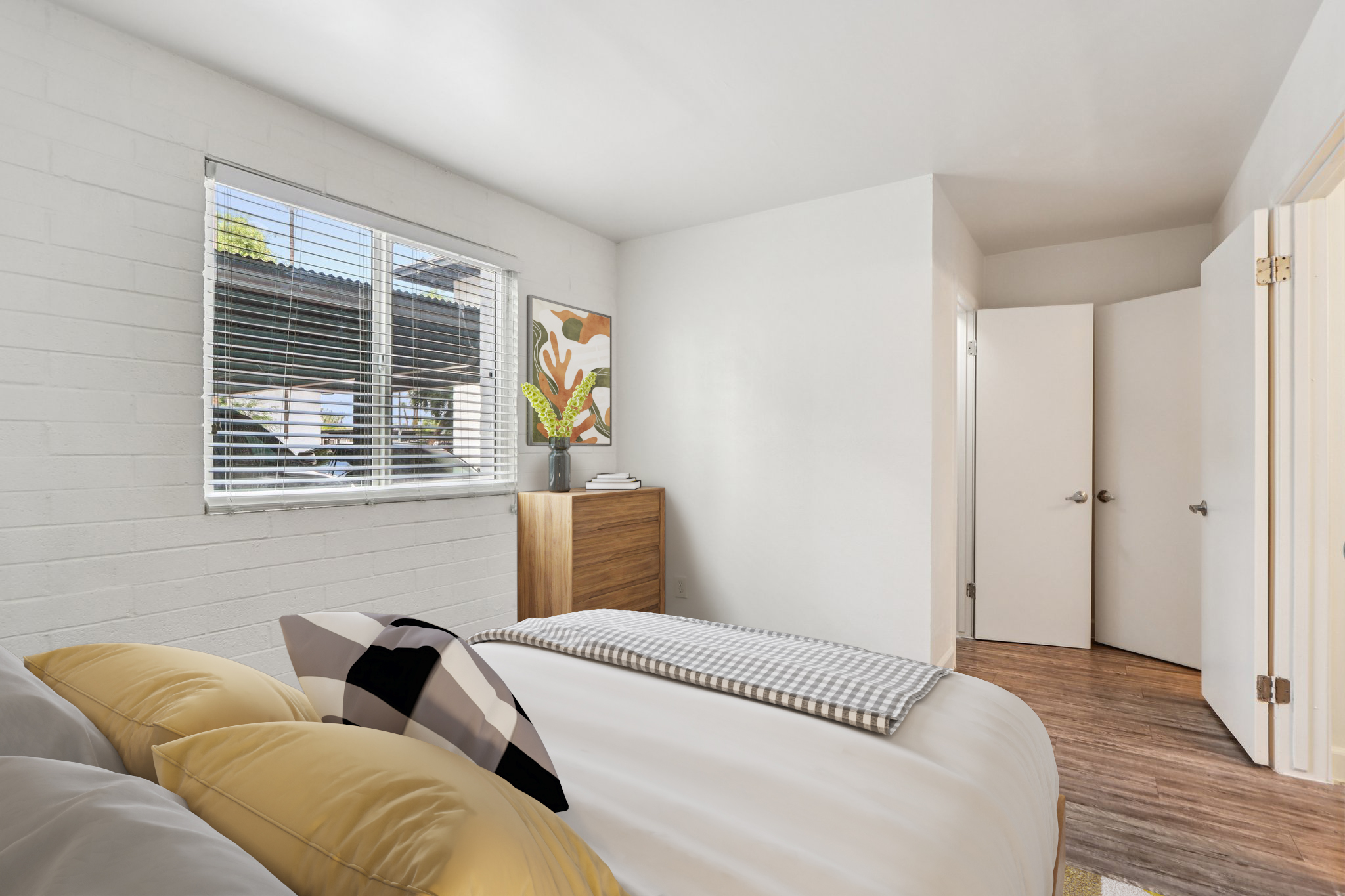 Cozy bedroom featuring a large bed with yellow and black pillows, a modern wooden dresser, and a colorful artwork on the wall. Natural light enters through a window with blinds, illuminating the minimalist decor and welcoming atmosphere. The door leads to another room, emphasizing the room's openness.