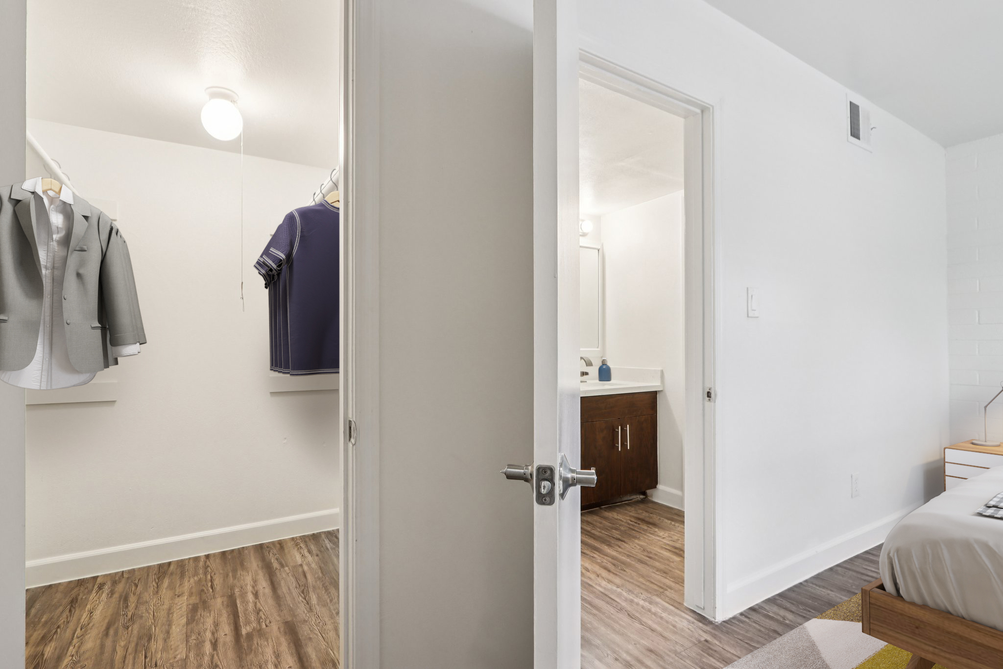 A well-lit interior of a modern apartment featuring an open door to a closet with hanging clothes, including a gray suit and a purple shirt. Adjacent to the closet, a bathroom is visible with a vanity mirror and white cabinetry, while a cozy bedroom with a bed is also in view, showcasing hardwood flooring.
