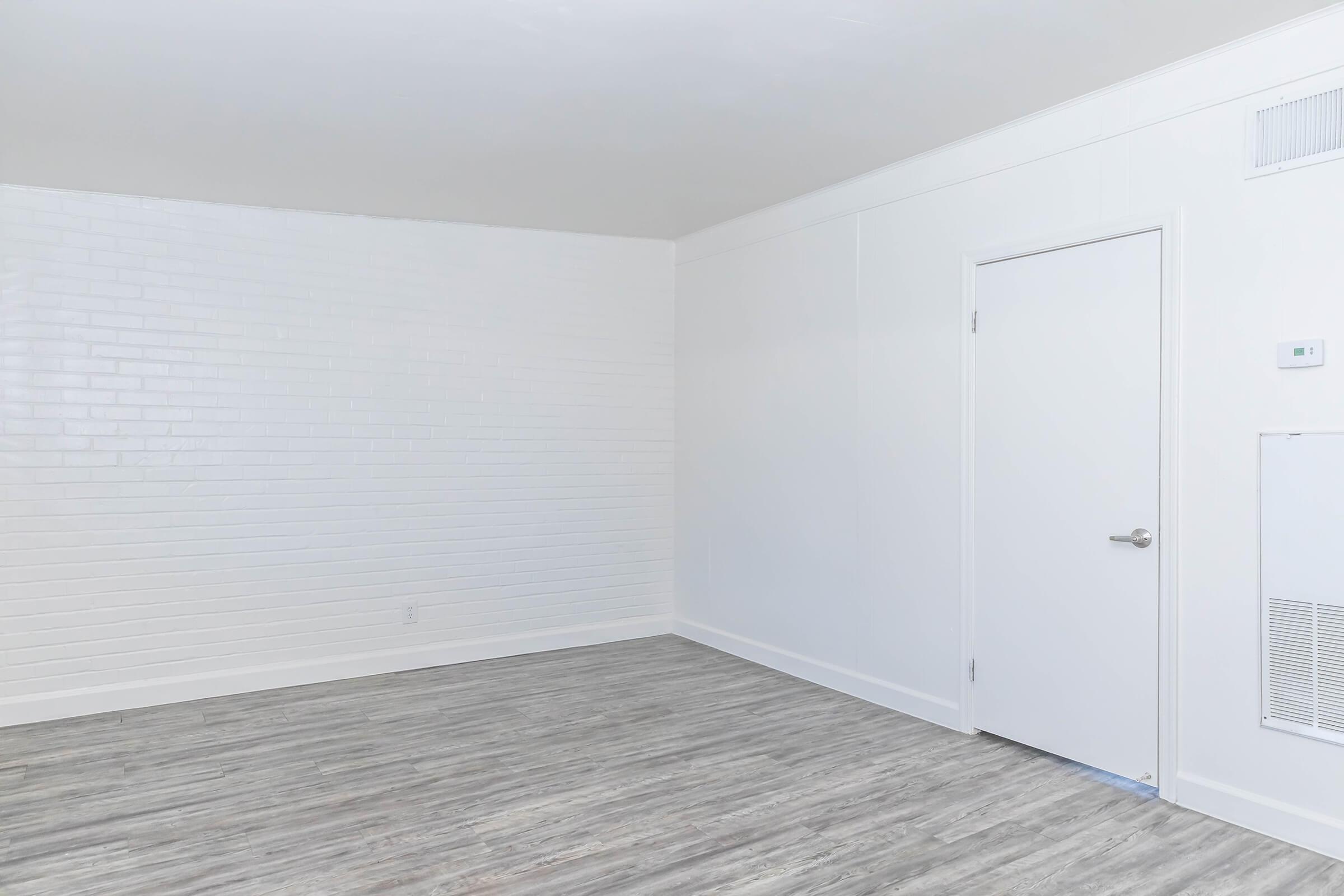 Empty room with a white brick wall and a plain white door. The floor is light-colored laminate. The space appears clean and minimalistic, with no furniture or decoration present.