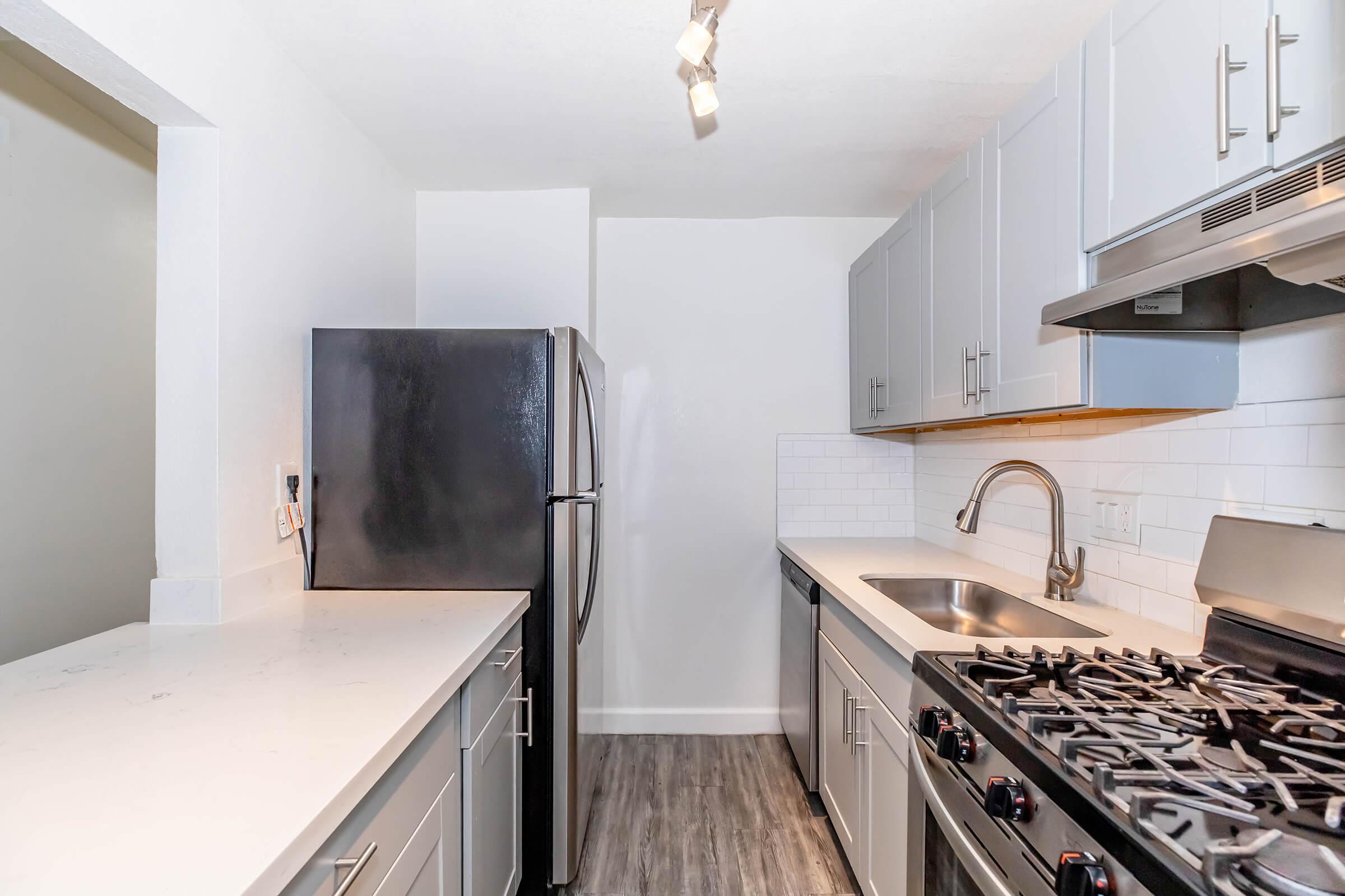 A modern kitchen featuring light gray cabinets, a stainless steel sink, and a black refrigerator. The kitchen includes a gas stove with multiple burners and a sleek countertop. Bright lighting enhances the spacious appearance, with white tile backsplash adding a clean look.