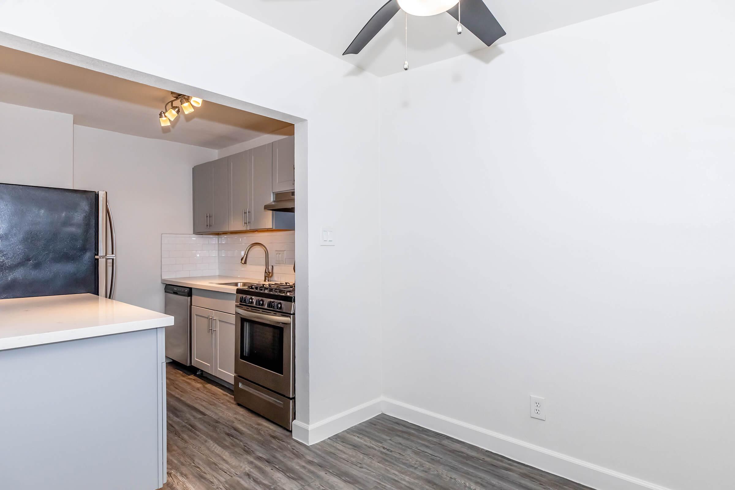 A modern kitchen featuring stainless steel appliances, white cabinetry, and a sleek countertop. There is a ceiling fan visible, and the space has light wood-style flooring. The layout includes a connected cooking and dining area with a minimalistic design and ample natural light.