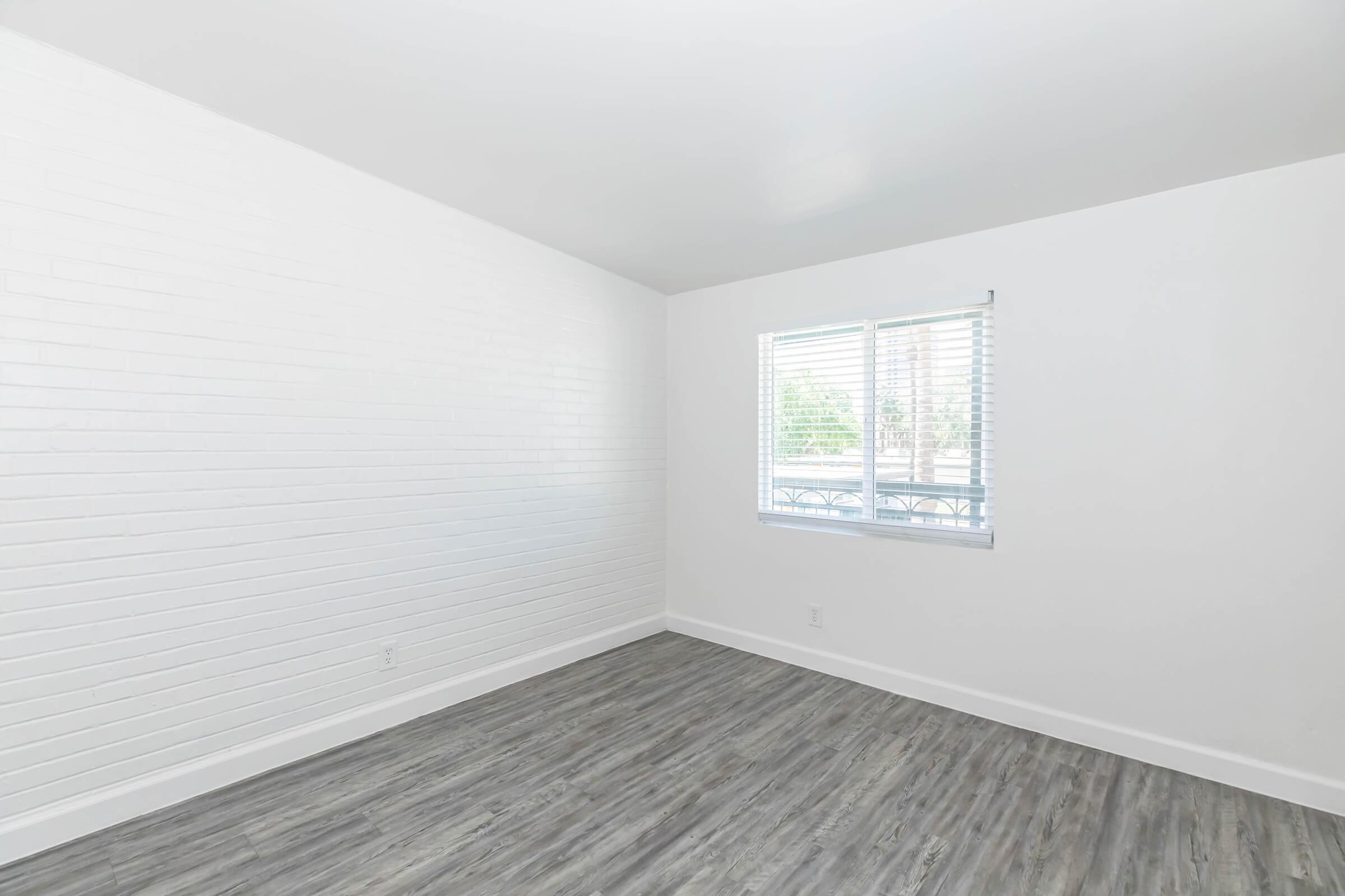 Empty room with white brick walls and light-colored wood flooring. A window with horizontal blinds allows natural light to enter, illuminating the space. The overall aesthetic is modern and minimalistic, creating a sense of openness.