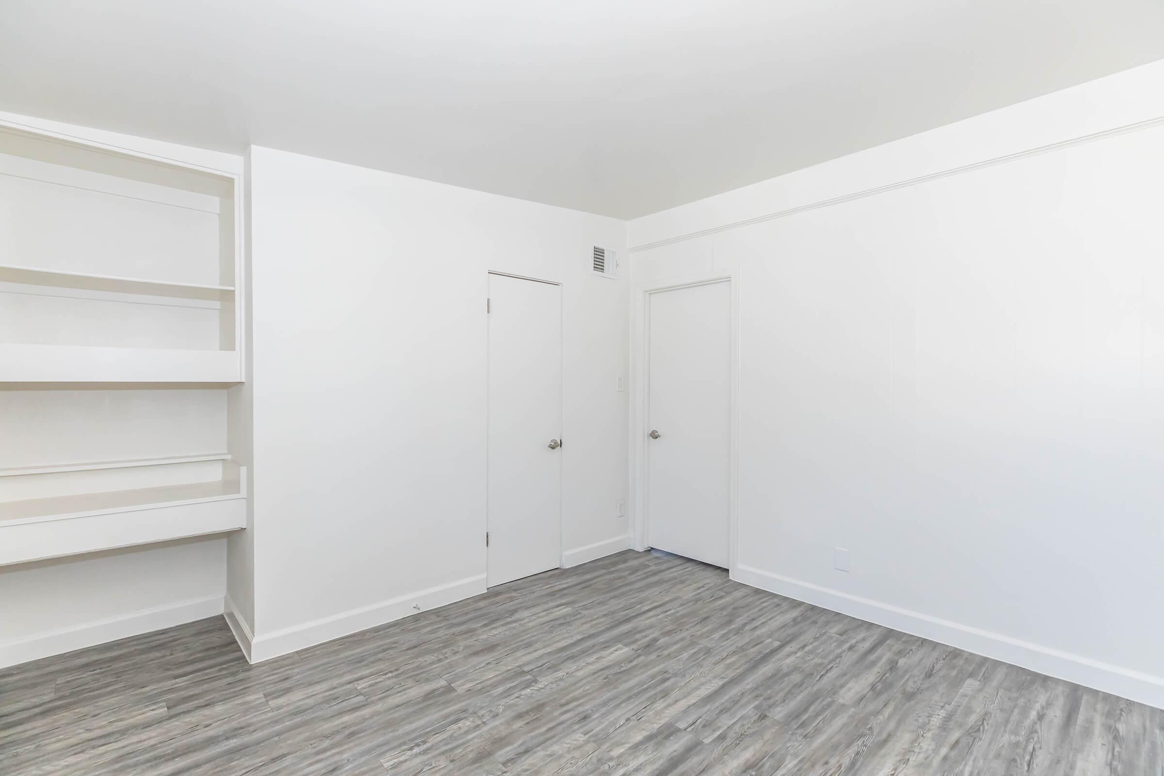 A minimal, empty room with light-colored walls and a smooth, gray wooden floor. It features a built-in shelf on the left, a closed door on the right, and another door opposite the shelf, creating a simple and clean aesthetic. Natural light may enhance the spacious feel of the area.