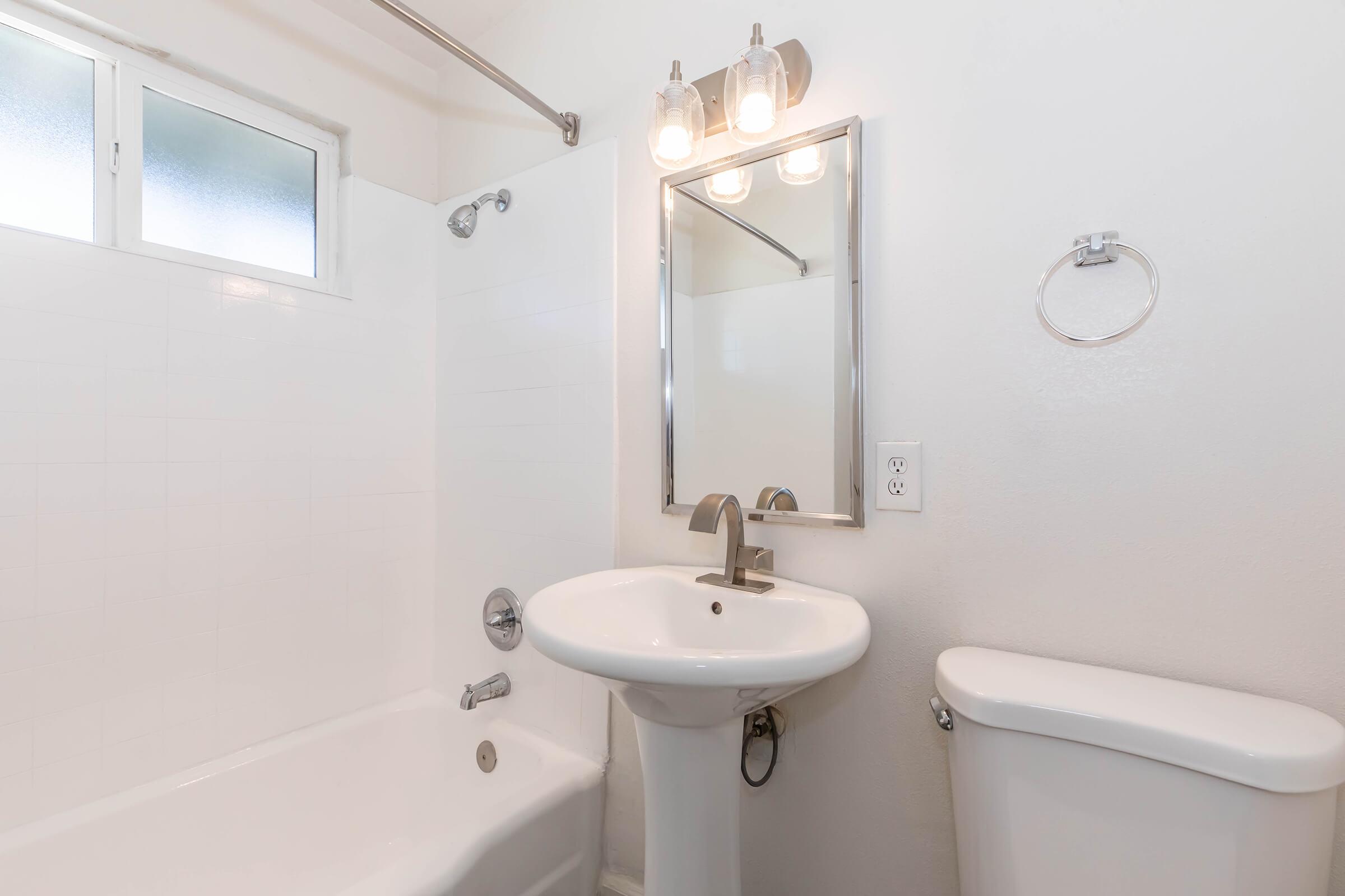 A clean, modern bathroom featuring a white bathtub with a shower, a pedestal sink, a large wall mirror, and a towel holder. The walls are painted light colors, and there's a window allowing natural light to enter, enhancing the space's brightness.