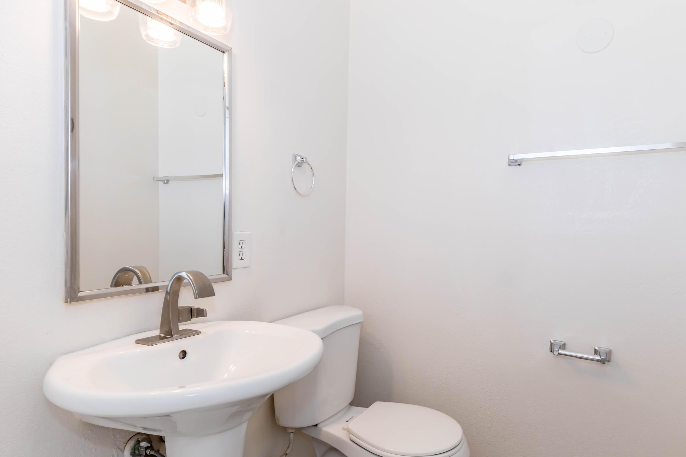 A clean, minimalistic bathroom featuring a white toilet and a pedestal sink with a modern faucet. A rectangular mirror is mounted above the sink, and a towel bar is attached to the wall next to it. The walls are painted white, enhancing the bright and airy feel of the space.