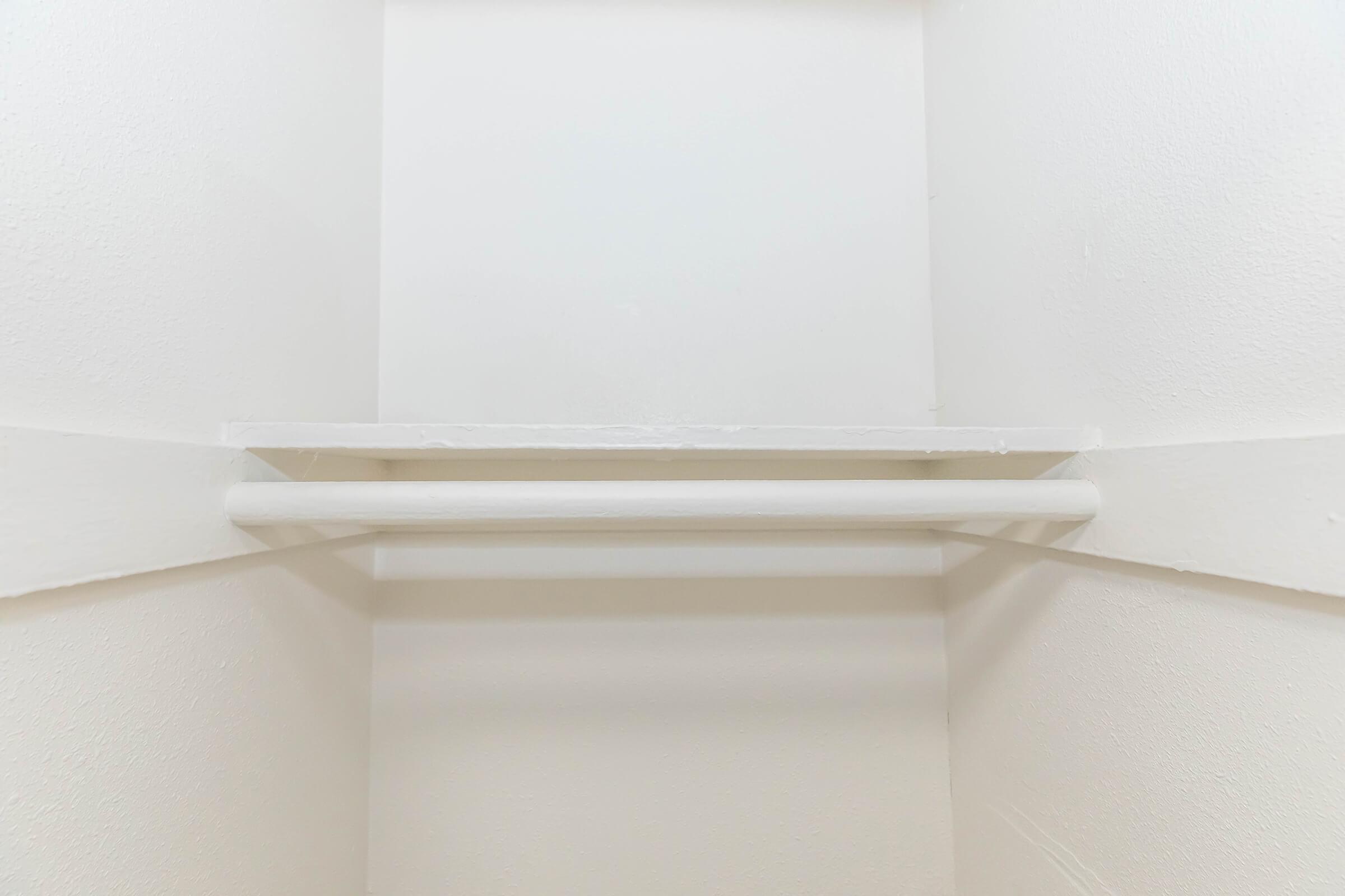 A minimalist, empty closet with white walls. A narrow, white shelf is mounted against the wall above a horizontal white rod, typically used for hanging clothes. The overall look is clean and simple, emphasizing a bright and spacious feel.