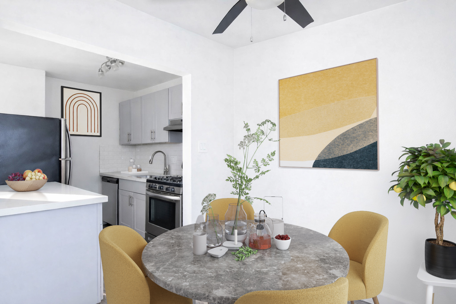 Modern kitchen and dining area featuring a round table with yellow chairs, a bowl of fruit, and decorative plants. The kitchen has gray cabinets, a stainless steel stove, and a black refrigerator. A large abstract artwork hangs on the wall, and there’s a ceiling fan above. Bright, minimalist design with light colors.