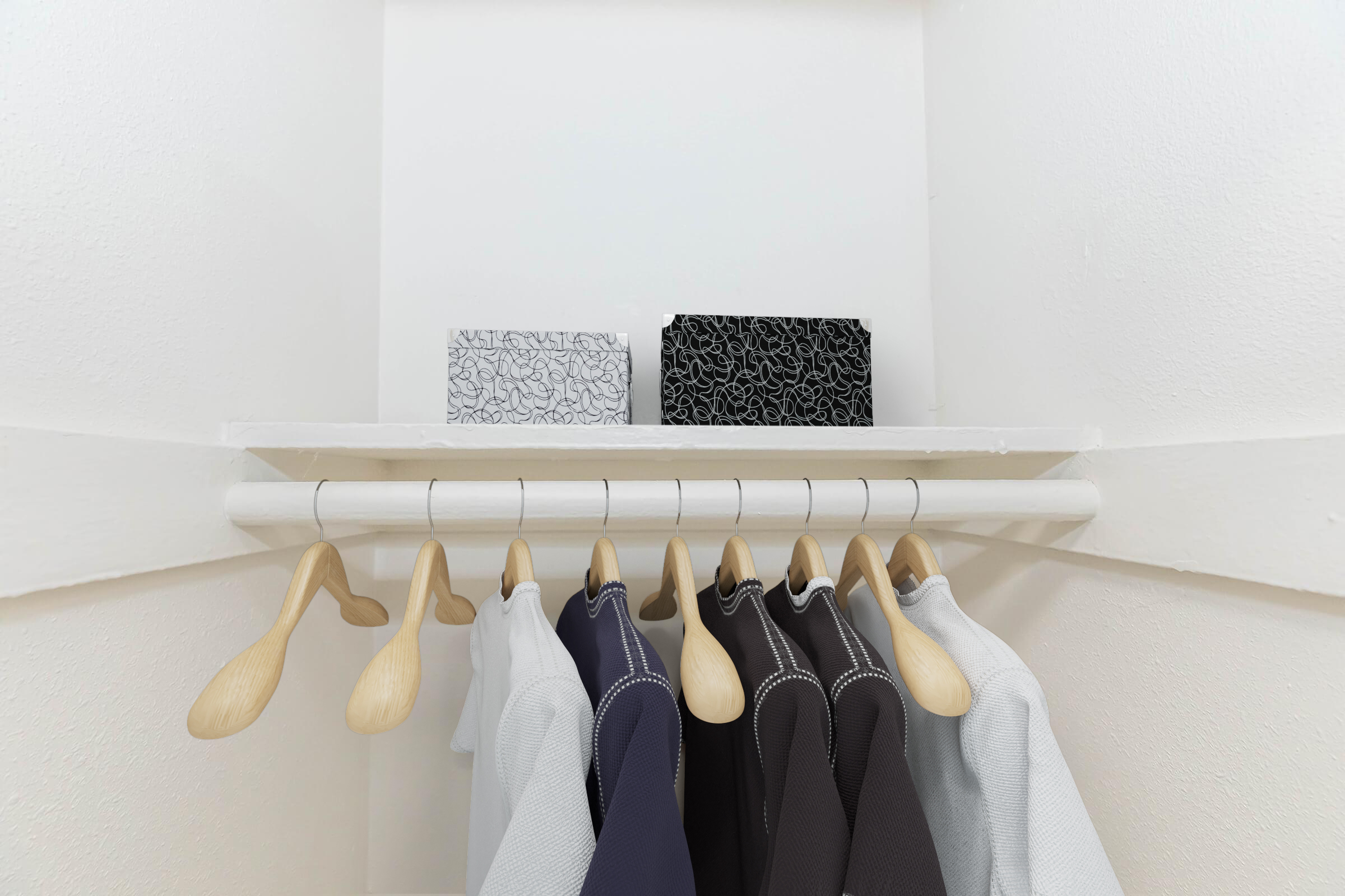 A neatly organized closet shelf with six hanging garments on wooden hangers. Above the clothes, there are two boxes with intricate black and white patterns. The closet has a clean, minimalist appearance with light-colored walls.